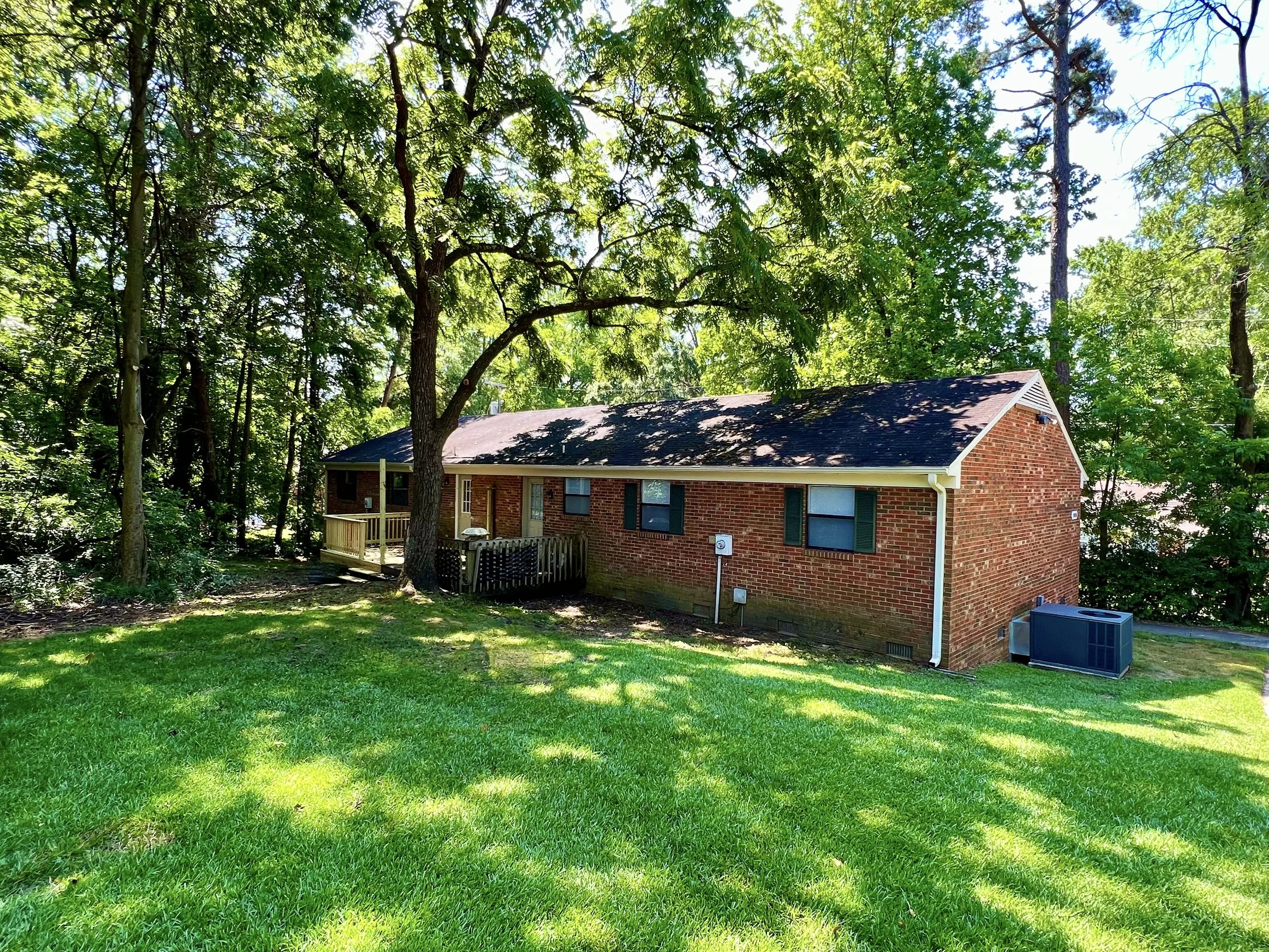 A backyard with green grass, a large tree, and a brick house with a porch and a small fenced yard, under bright sunlight.