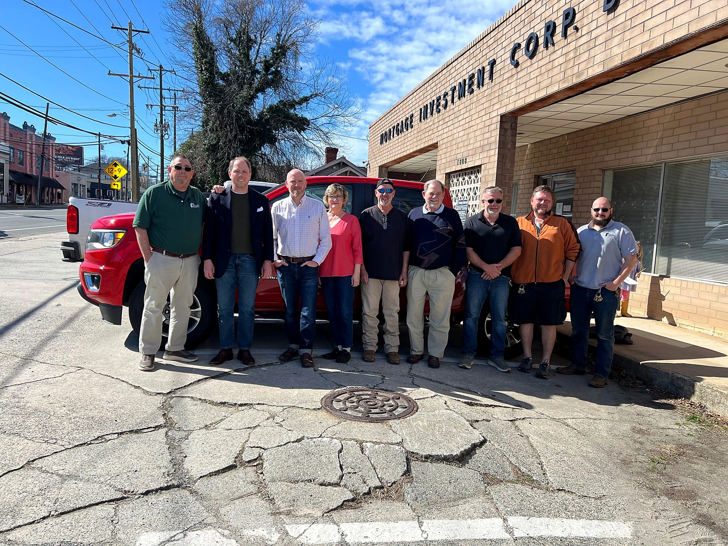 A group of nine people standing in front of a brick building with the sign 'Veterans Investment Corp' on it, on a sunny day with a blue sky.