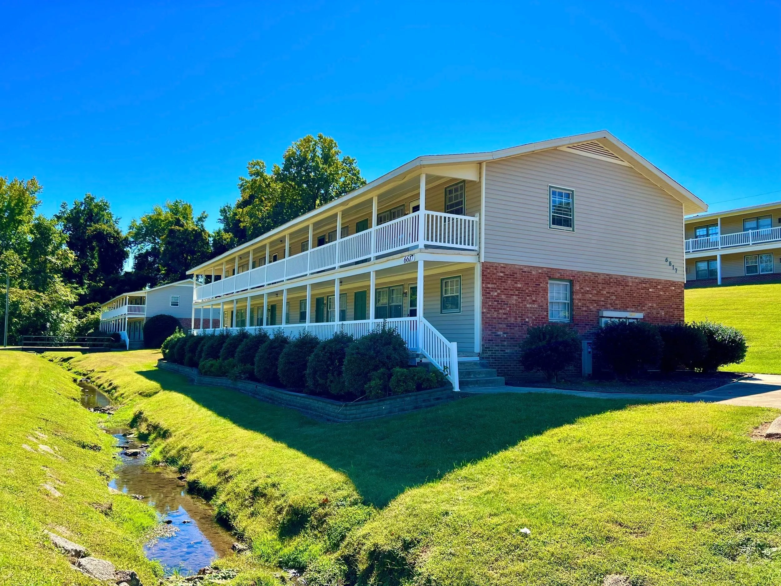 A multi-story residential building with beige siding, white balconies, and brick foundation, situated on well-maintained green lawn with landscaped bushes and a small creek nearby, under a bright blue sky.