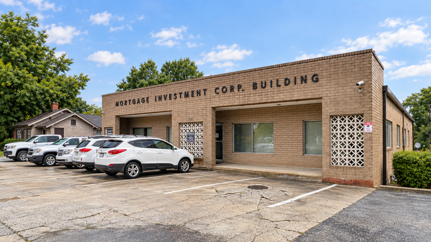 A brick building labeled 'Mortgage Investment Corp. Building' with a parking lot in front containing several parked cars and surrounded by trees under a partly cloudy sky.
