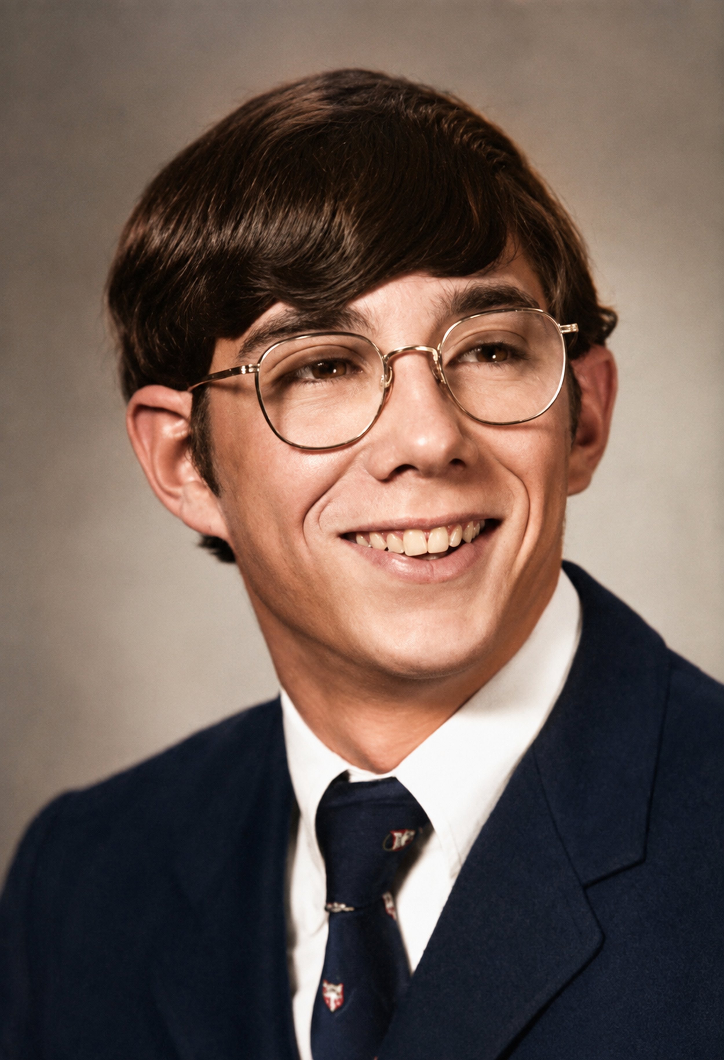 Portrait photograph of a young man with brown hair, glasses, wearing a dark suit, white shirt, and patterned tie, smiling.