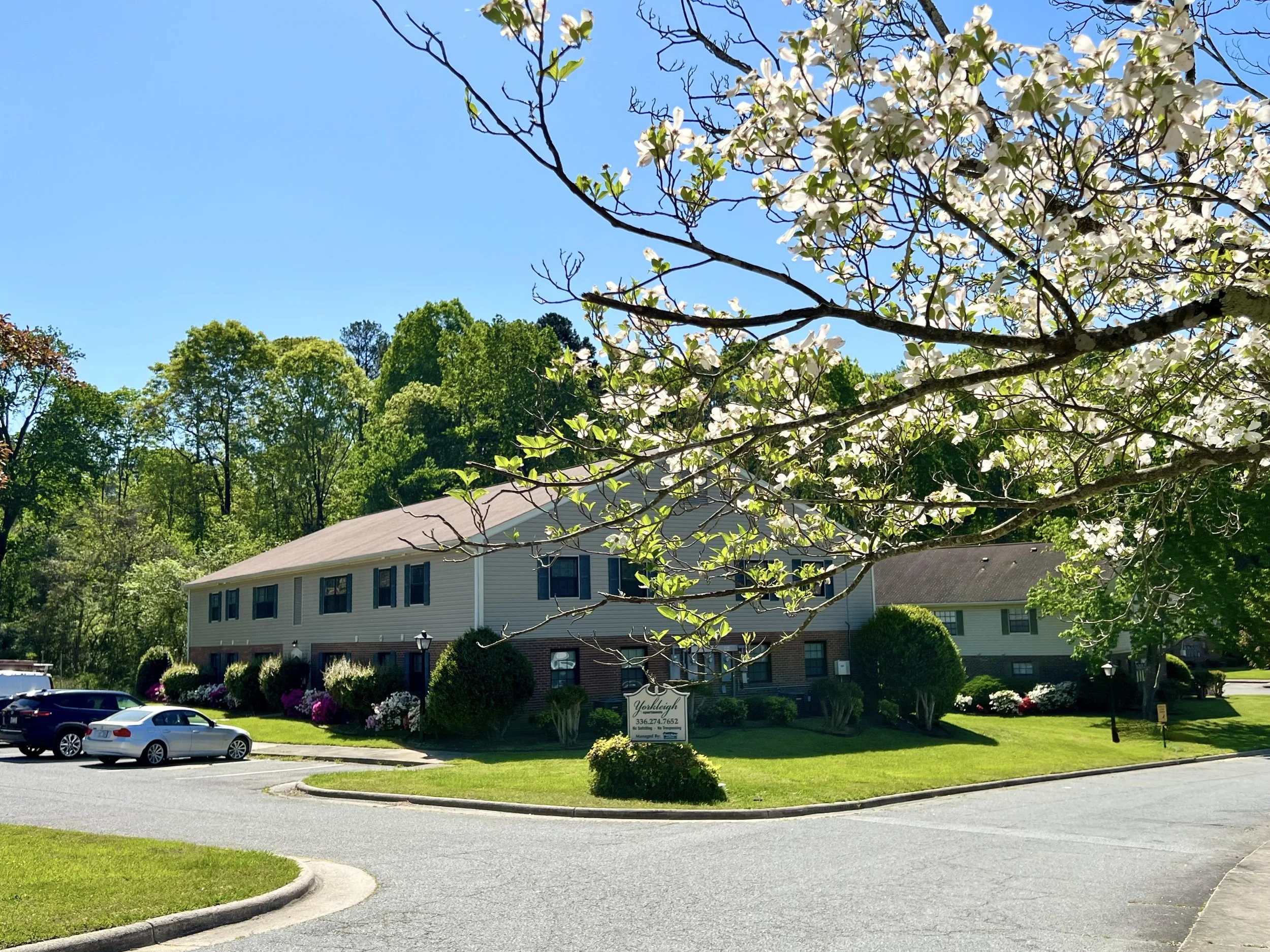 A residential apartment complex surrounded by a well-maintained lawn, flowering bushes, and several parked cars under a clear blue sky. There is a white building with multiple windows and a sign that reads 'Yorlelight' near the entrance.