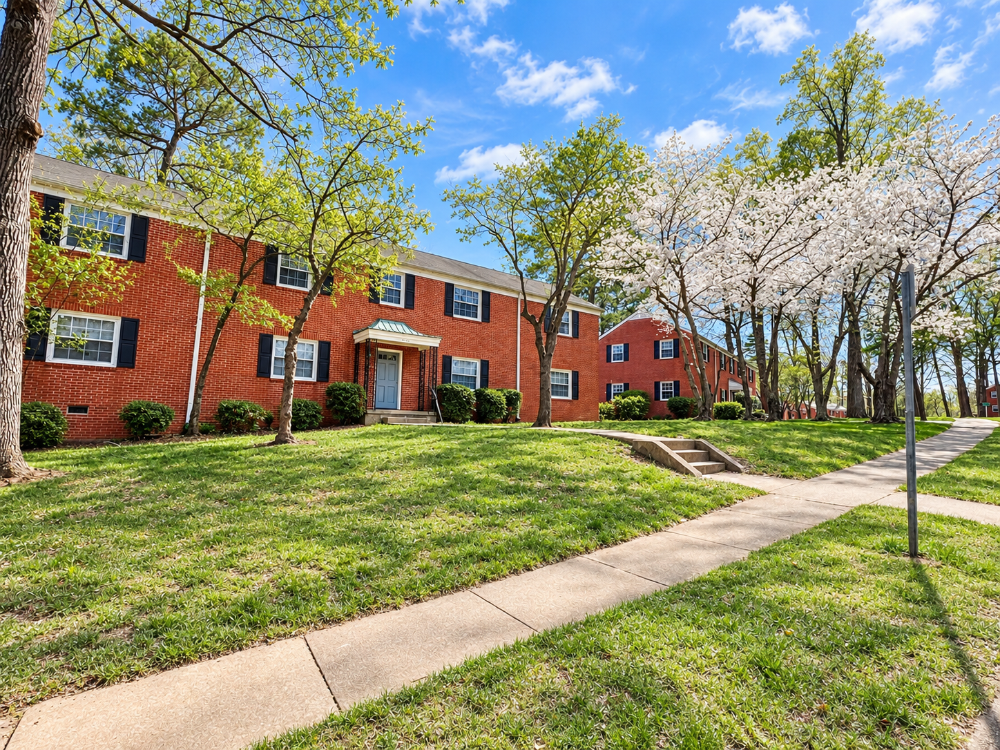 Red-brick apartment building with black shutters, surrounded by green grass and trees with new leaves and white blossoms, under a blue sky with some clouds.