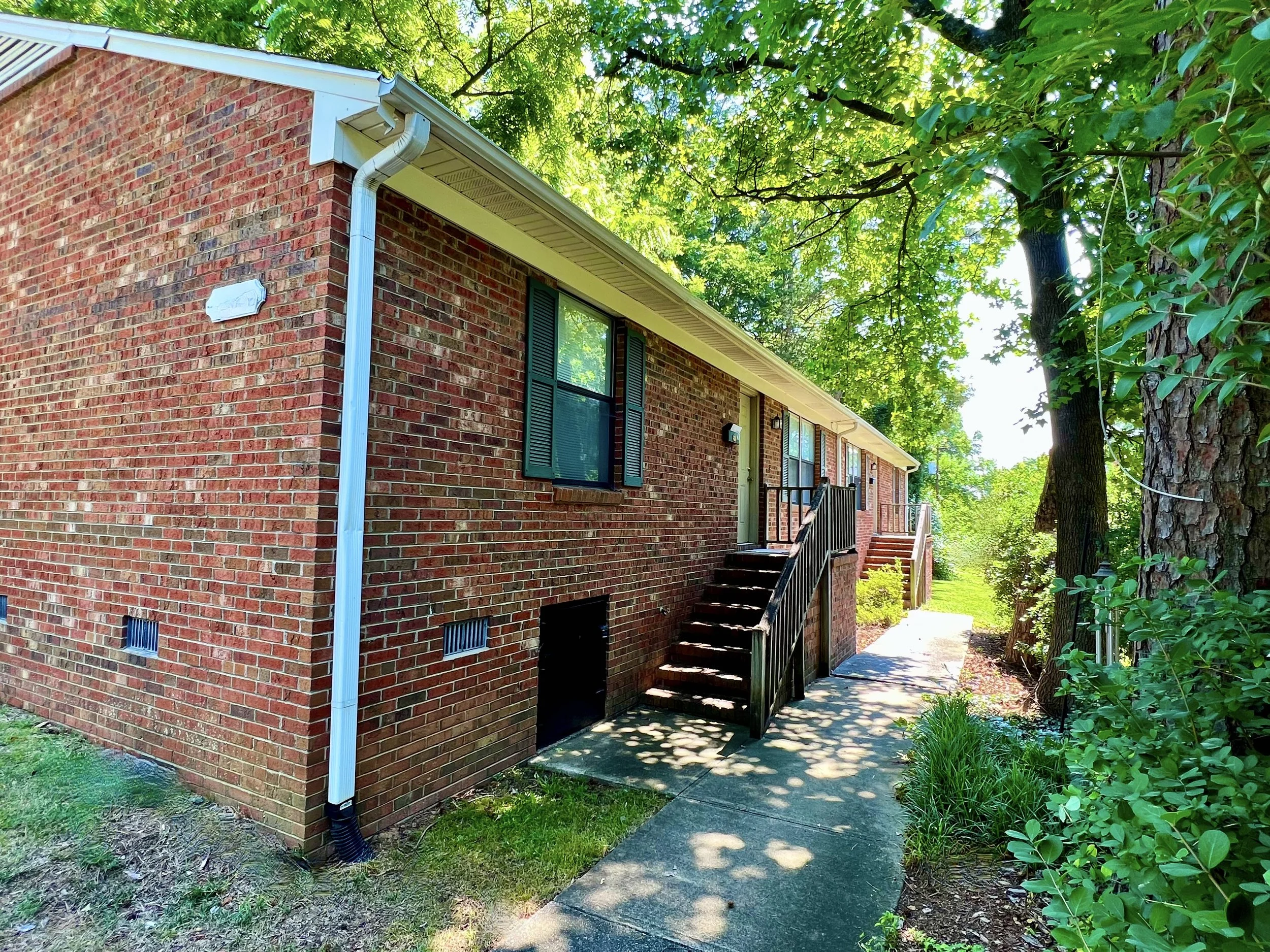 A brick residential building with a staircase leading to a door, surrounded by green trees and bushes on a sunny day.