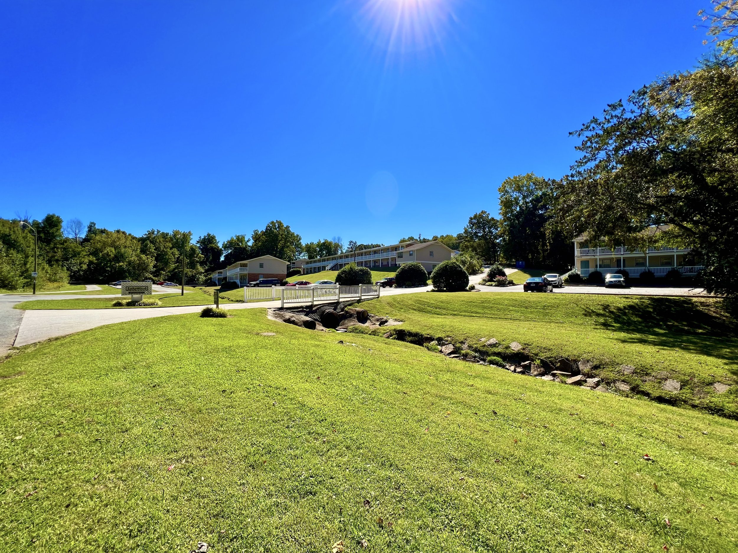 A sunny day in a suburban neighborhood with green lawns, trees, parked cars, and apartment buildings under a clear blue sky.