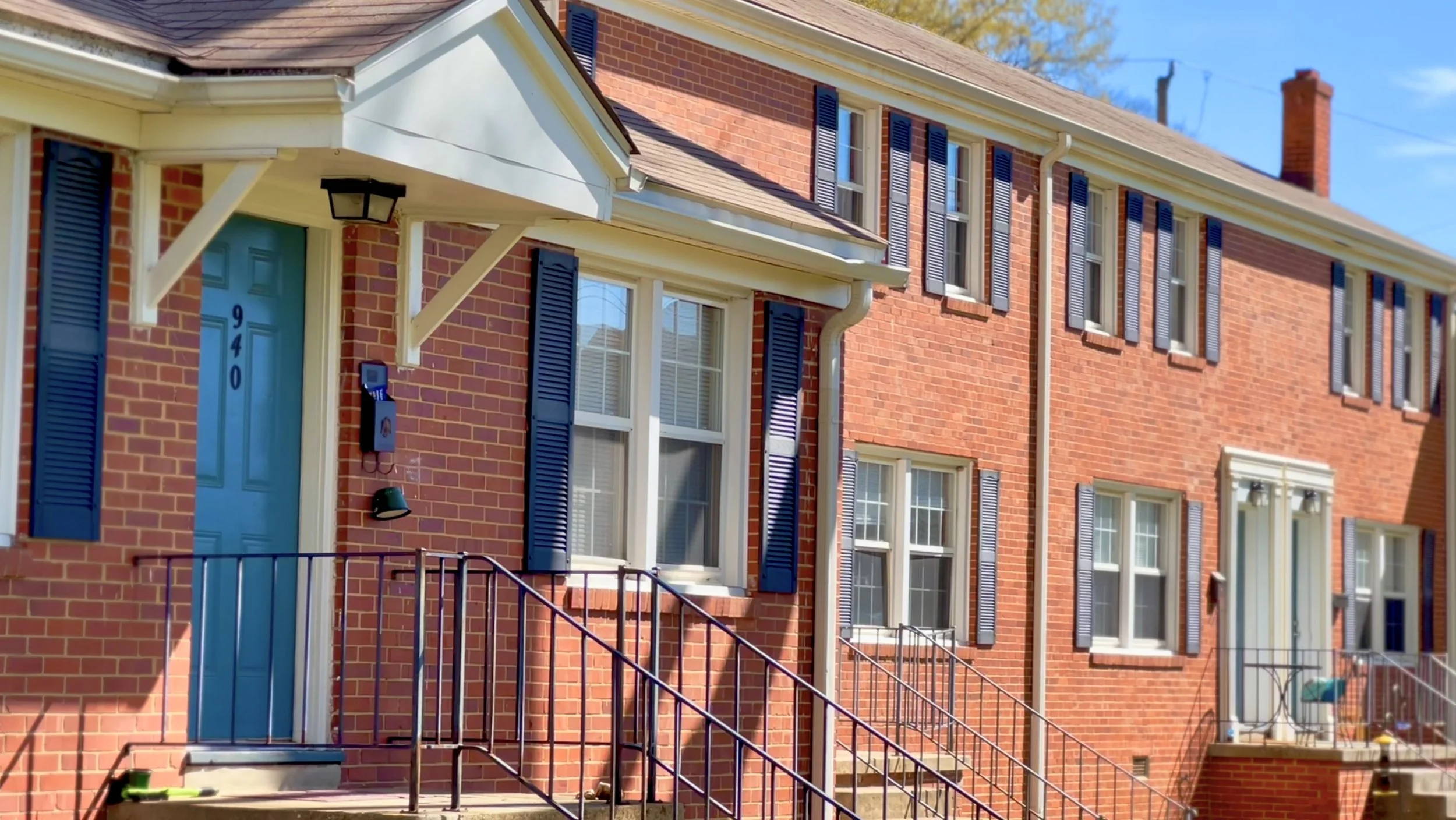 A row of brick townhouses with white trim, black shutters, and blue doors, featuring stairs and metal railings leading to the entrances, under a clear blue sky.