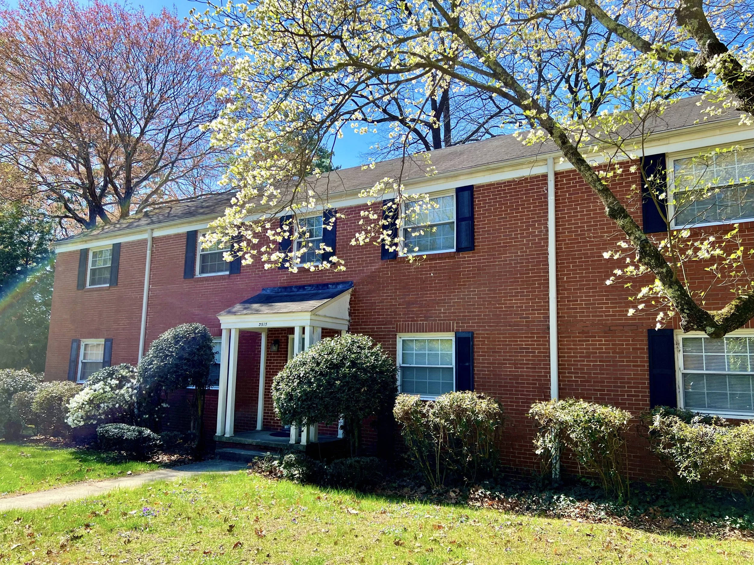A red brick two-story house with a white front porch, surrounded by trees and bushes, under a clear blue sky.