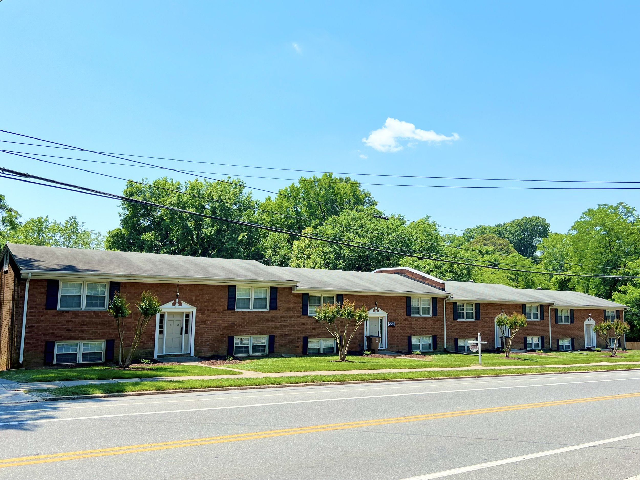 A row of brick apartment buildings with white doors and windows, small landscaped yards, and trees in front. Overhead power lines run across the scene under a clear blue sky with a small white cloud.