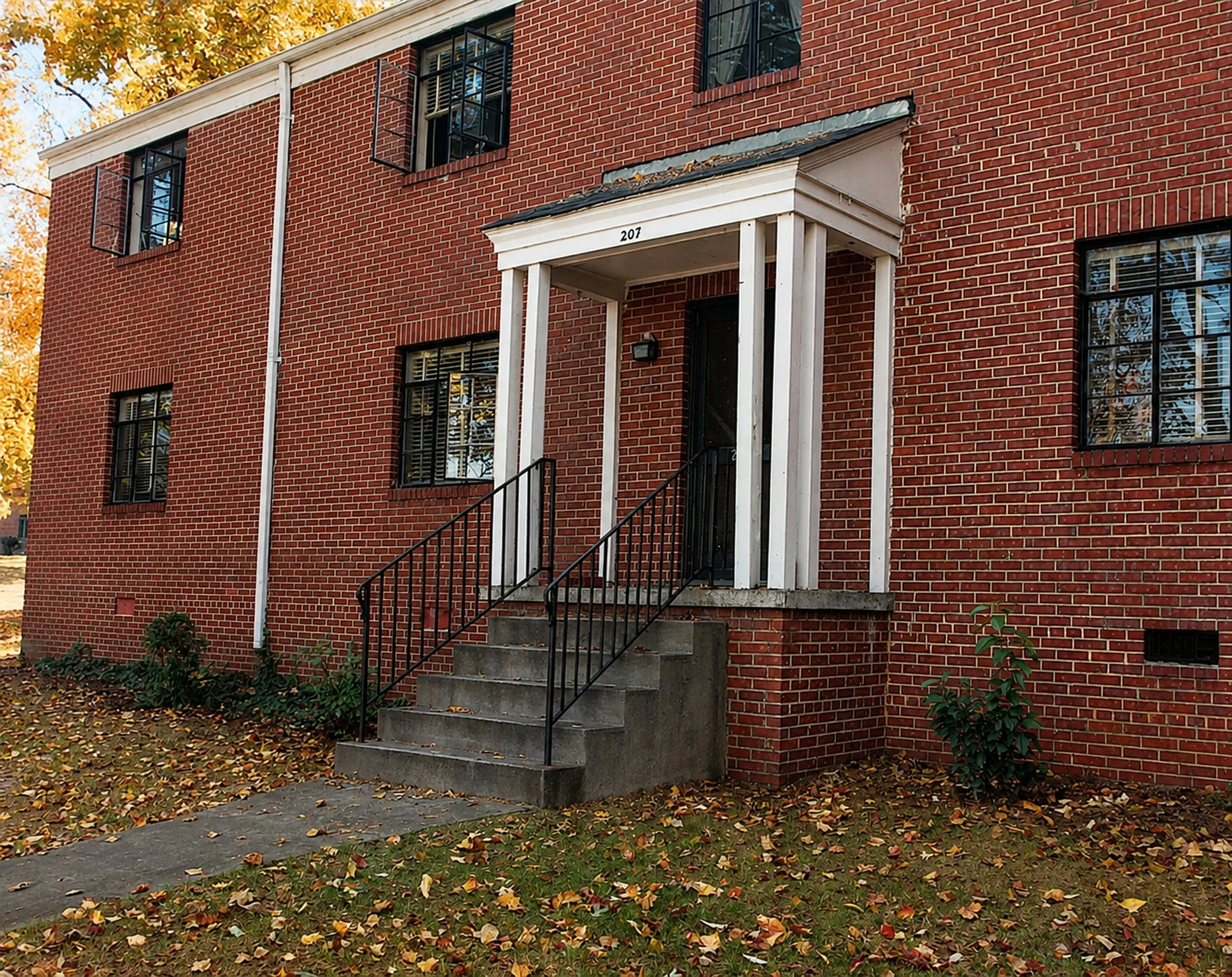 Brick apartment building with stairs leading to the front door, white trim, and open windows, surrounded by fallen autumn leaves.