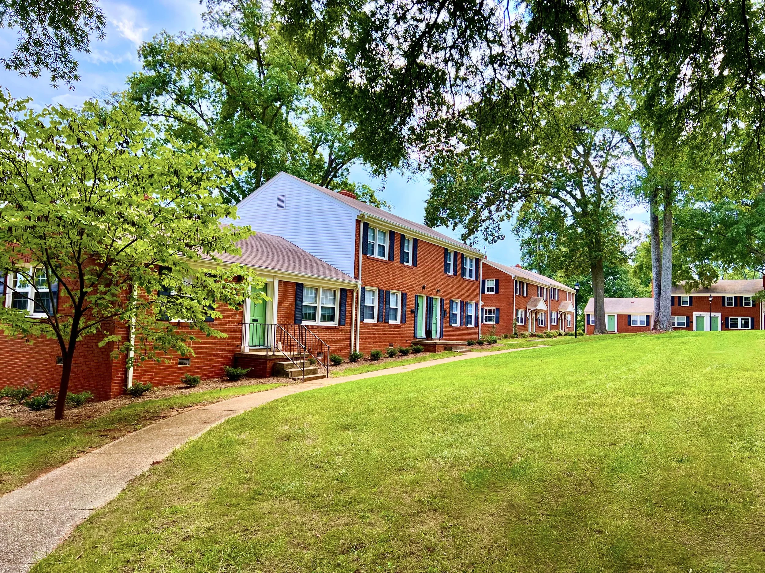 A row of residential townhouses with brick facades and green front doors, surrounded by lush green trees and a well-maintained grassy lawn, under a partly cloudy sky.
