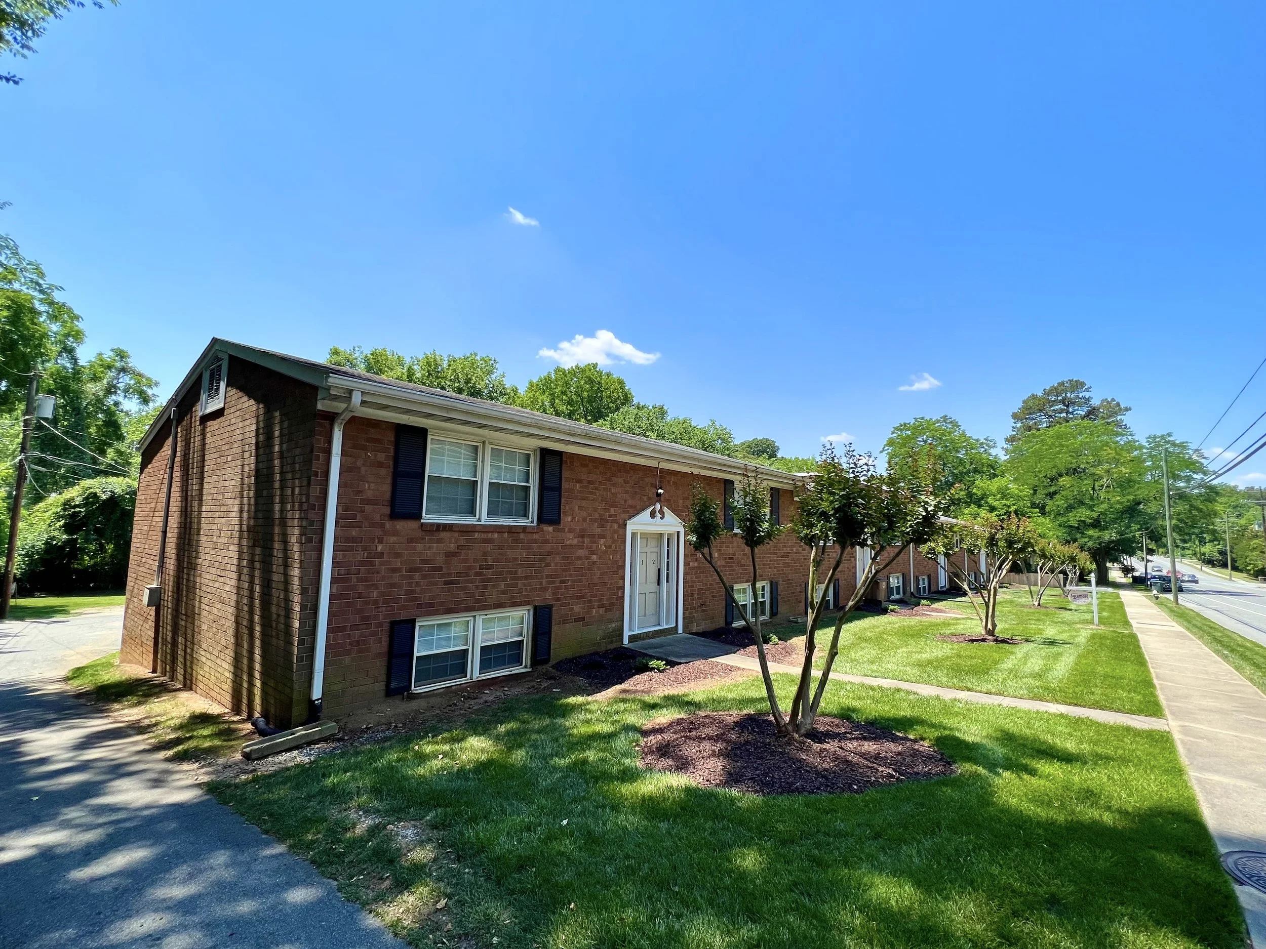 A brick apartment building with white windows and black shutters, surrounded by green trees and a well-maintained lawn, under a bright blue sky with a few clouds.
