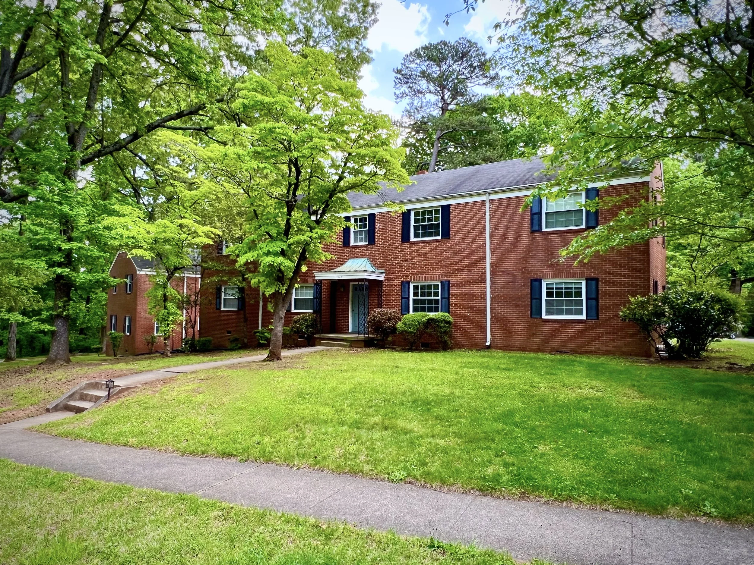 Front yard of a red-brick house with black shutters, surrounded by green trees and a well-kept lawn. A curved sidewalk leads to the house.