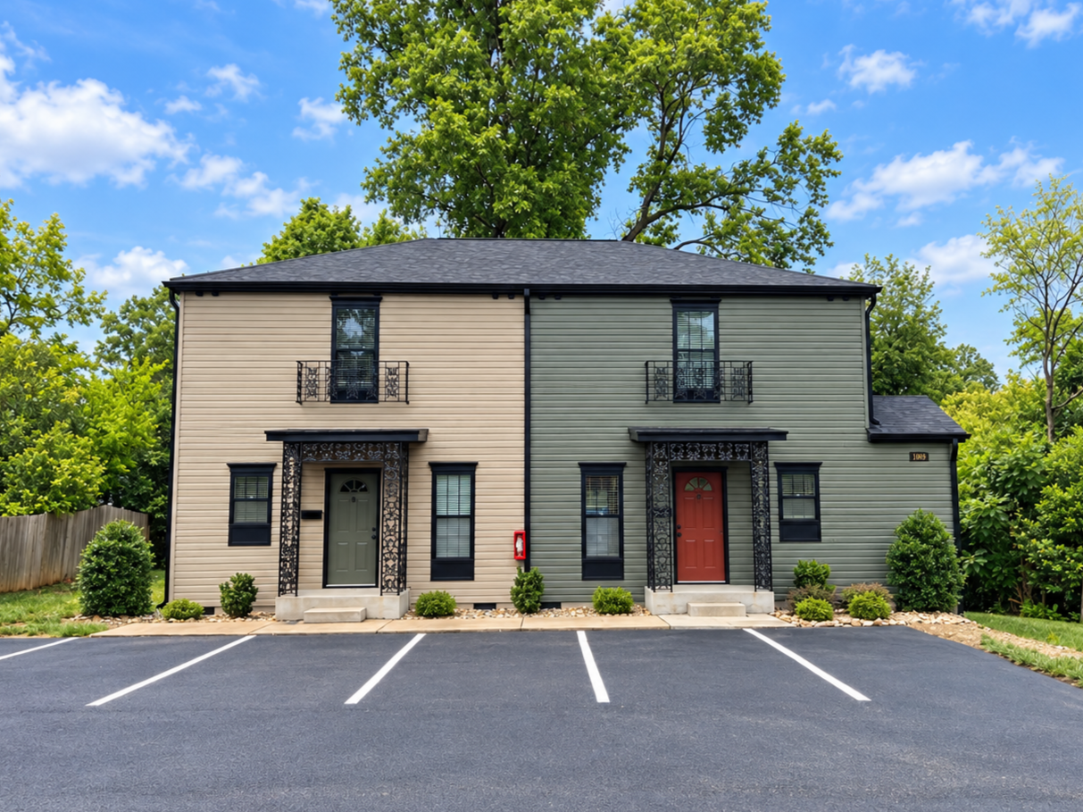 Two-story duplex with modern beige and green siding, black trim, and balconies on the upper level, fronted by a paved parking lot with white lines, small bushes, and trees in the background under a partly cloudy sky.