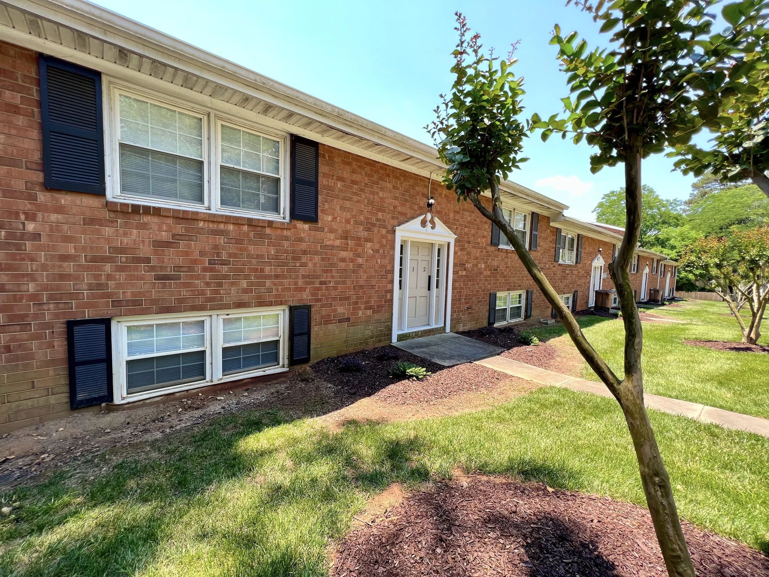 View of a red brick apartment building with white-framed windows with black shutters, white front door, and surrounding green lawn and trees under a clear blue sky.