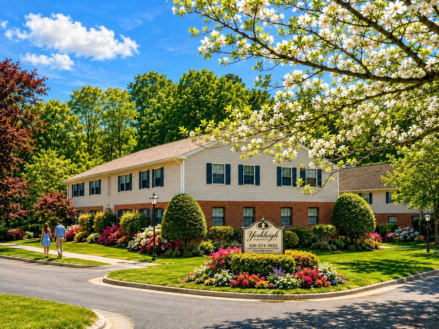 A residential apartment complex with a well-maintained lawn and colorful flower beds, a couple walking hand in hand, clear blue sky, and blooming trees.