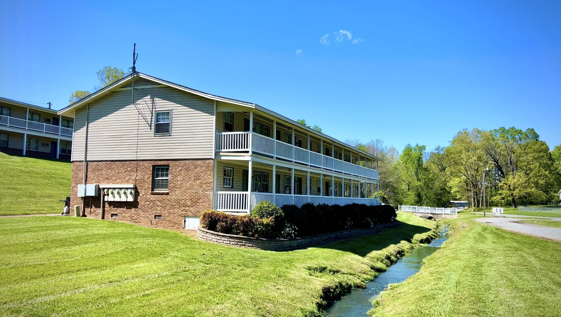 A multi-story residential building with white siding and a brick foundation, overlooking a landscaped yard and a small creek, under a clear blue sky.