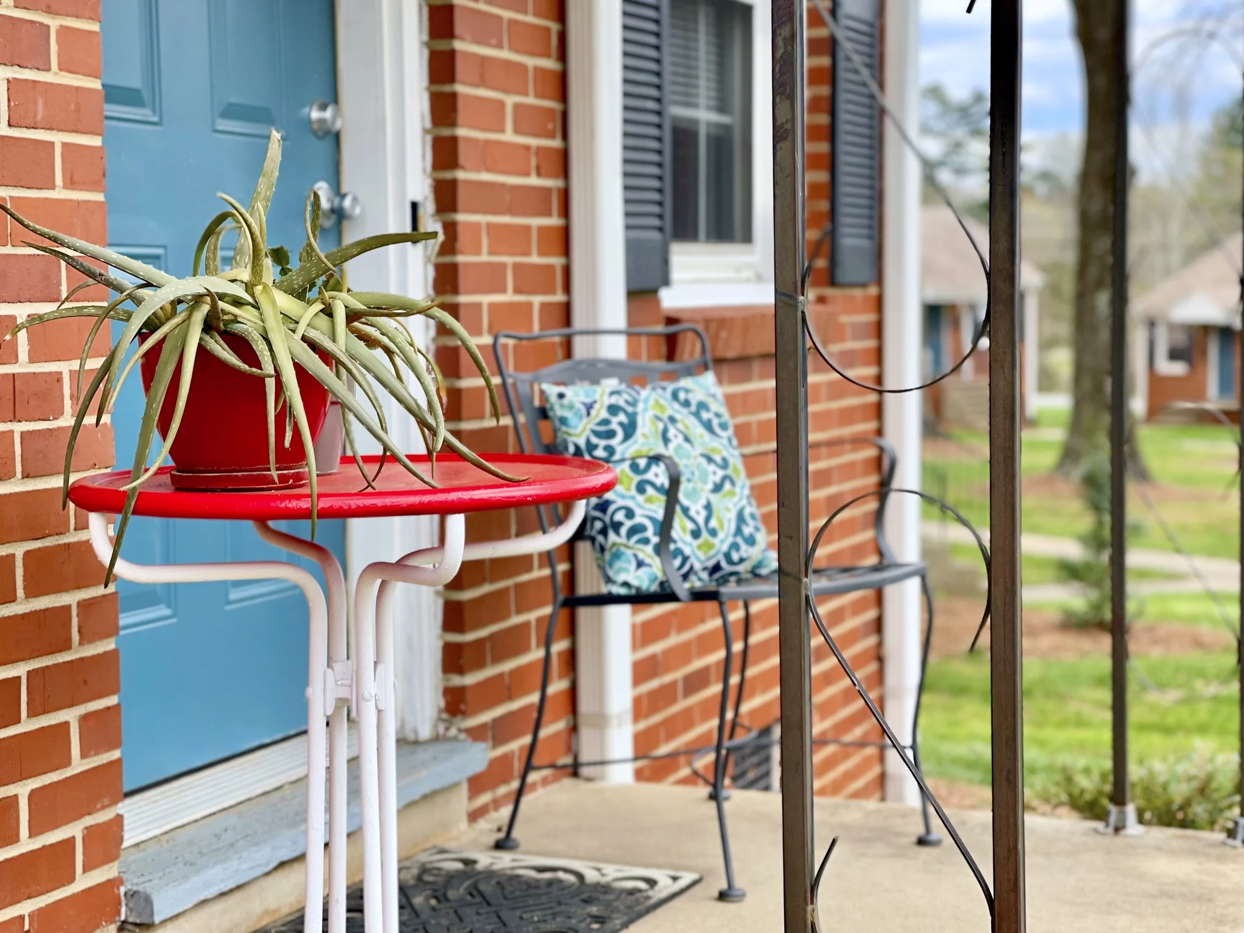 A porch scene featuring a red table with a potted aloe vera plant, a blue front door, a chair with a colorful cushion, and a black metal railing with a wire. The background shows part of a brick house with a window and black shutters, and some trees in the distance.