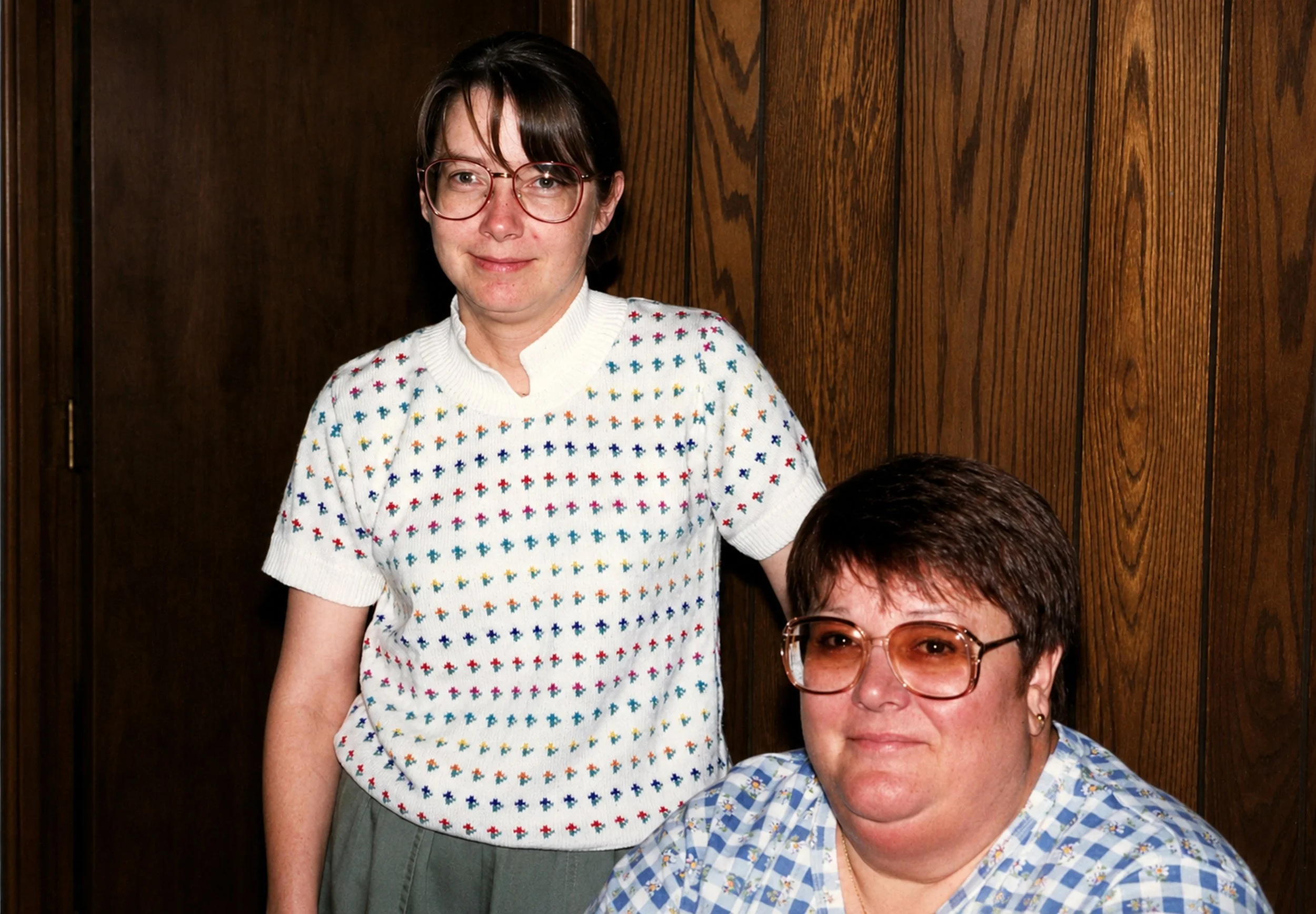 A woman with short brown hair and glasses wearing a white sweater with multicolored crosses, standing next to a woman with short dark hair, glasses, and a blue checkered blouse, both smiling in front of a wooden wall.