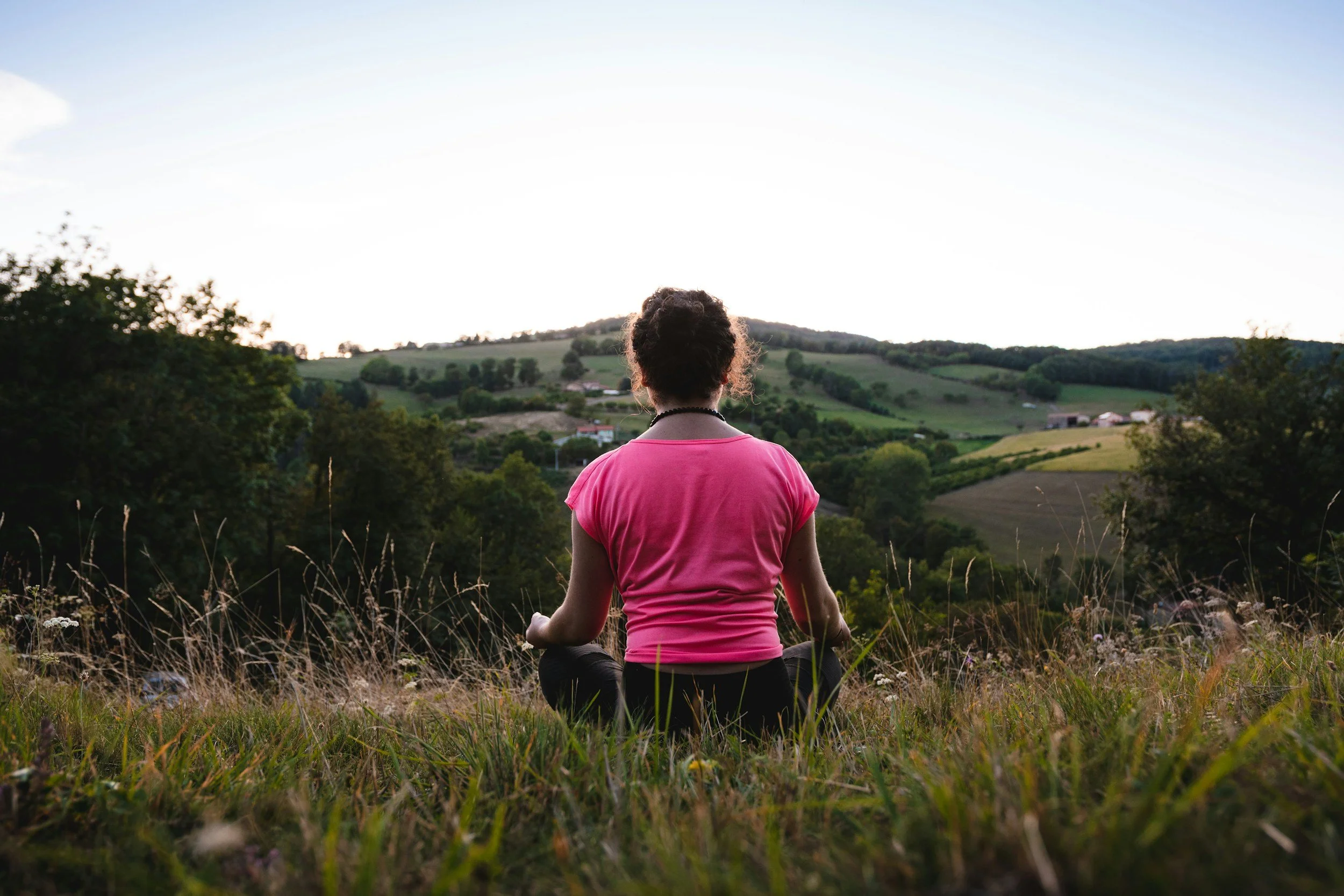 A person sitting in a meditative pose with crossed legs and arms resting on their knees, facing a scenic landscape of rolling green hills and scattered trees during sunset.