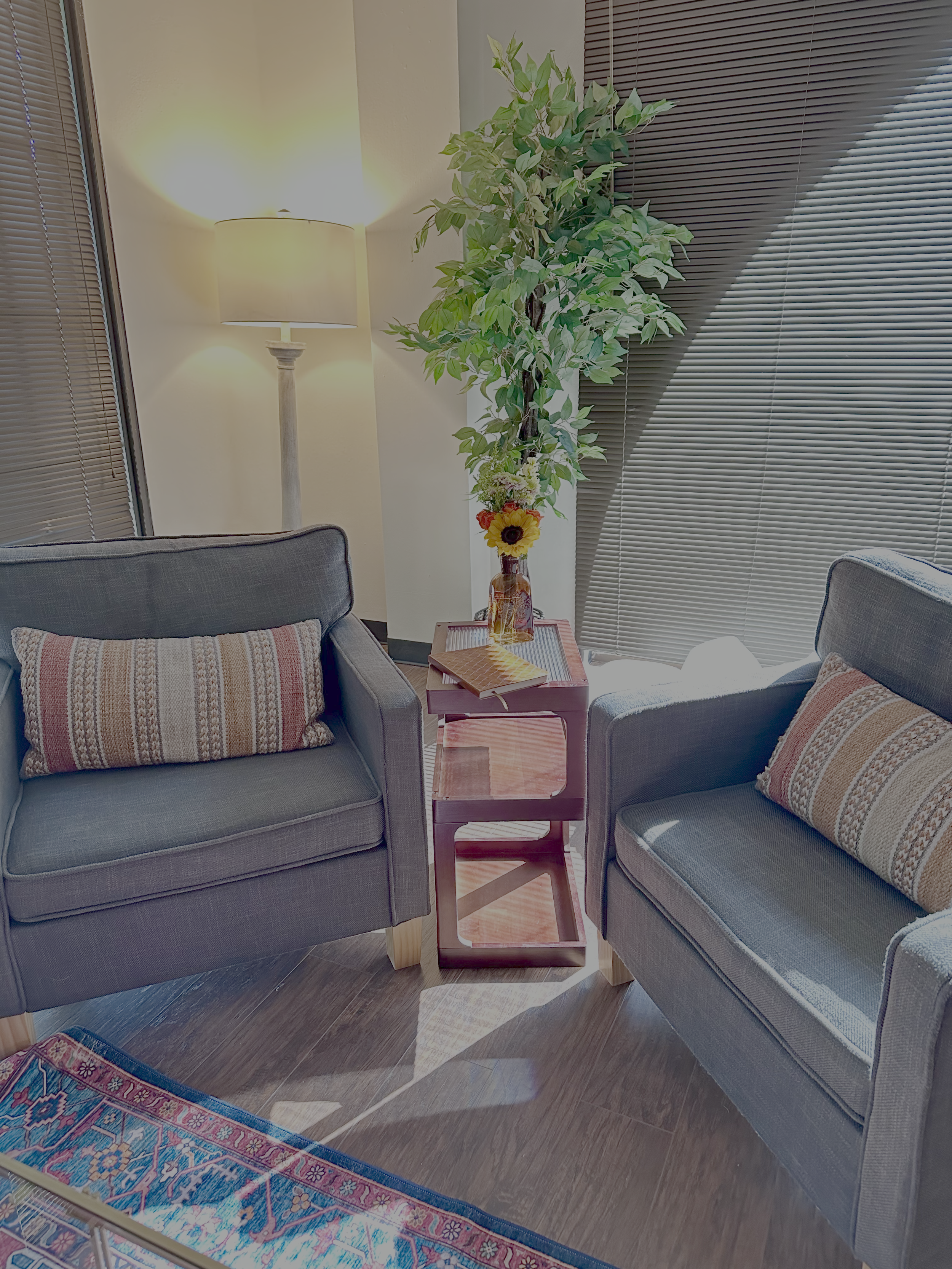 Counseling room with two gray upholstered armchairs, a small pink side table with a glass vase of flowers, a lamp, and window blinds.