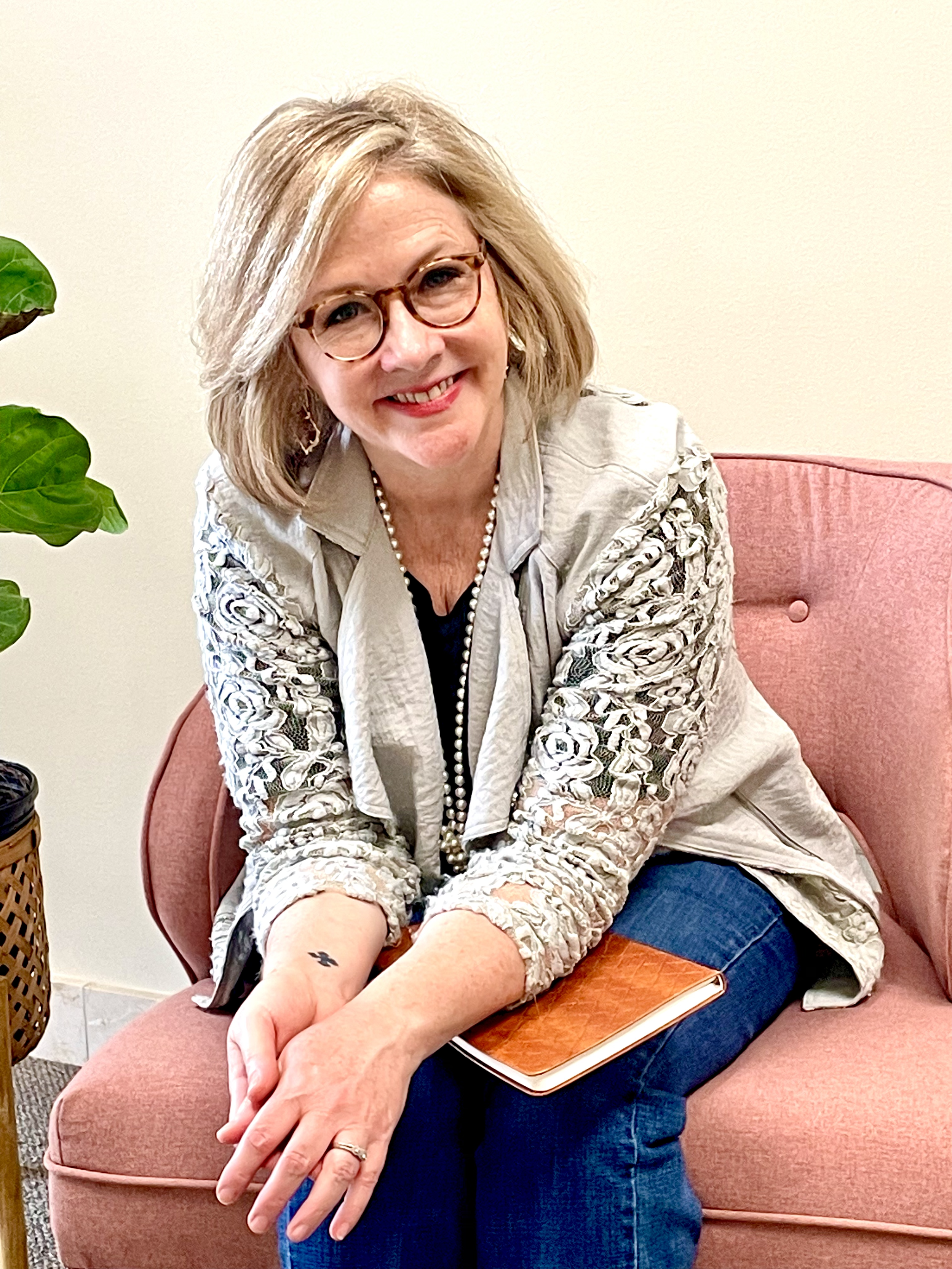 A woman with glasses and shoulder-length blonde hair, smiling and sitting on a pink couch, holding a notebook and wearing a beige jacket with embroidered sleeves and a pearl necklace.