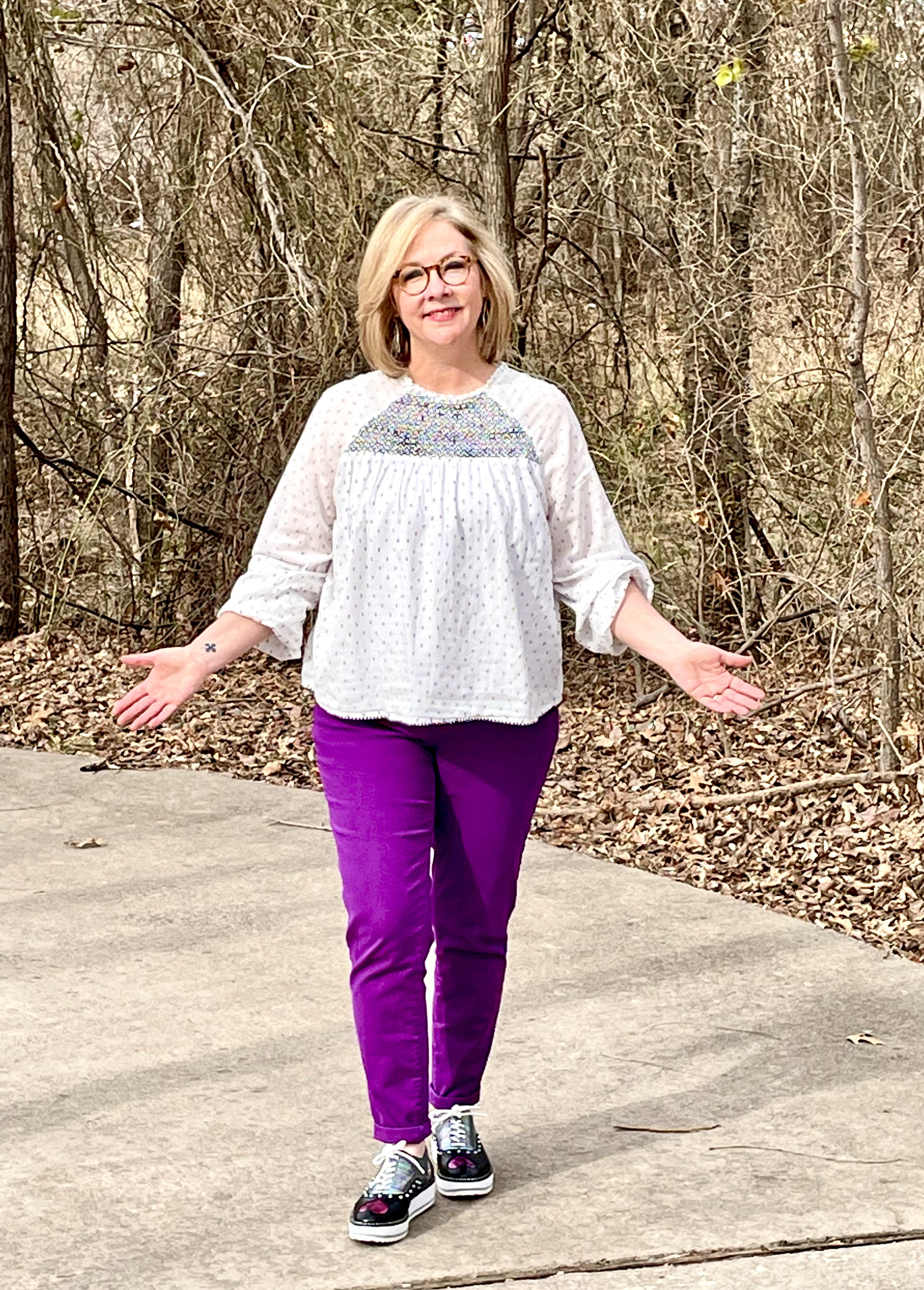 A woman with blonde hair and glasses standing outdoors on a concrete path, wearing a white blouse with purple pants and patterned sneakers, with a background of leafless trees and fallen leaves.