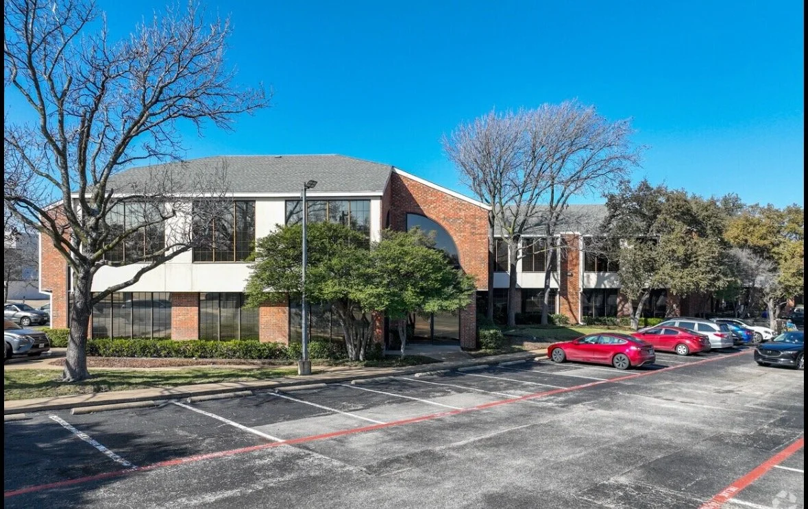 A parking lot in front of an office building with trees and parked cars, under a clear blue sky.