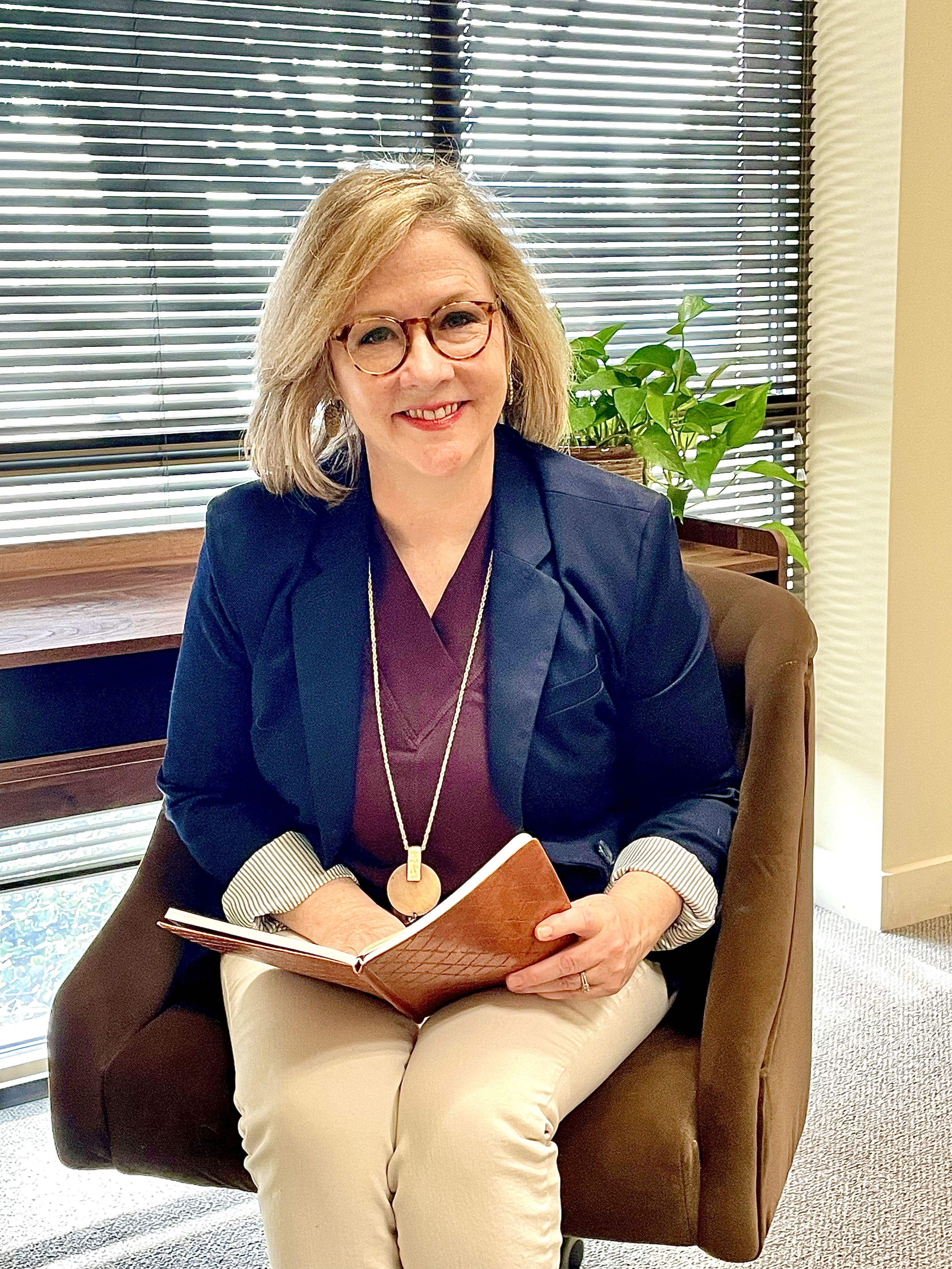 A woman with glasses and shoulder-length blonde hair sits in a leather chair, smiling, holding a notebook or journal, with a potted plant and window with blinds in the background.