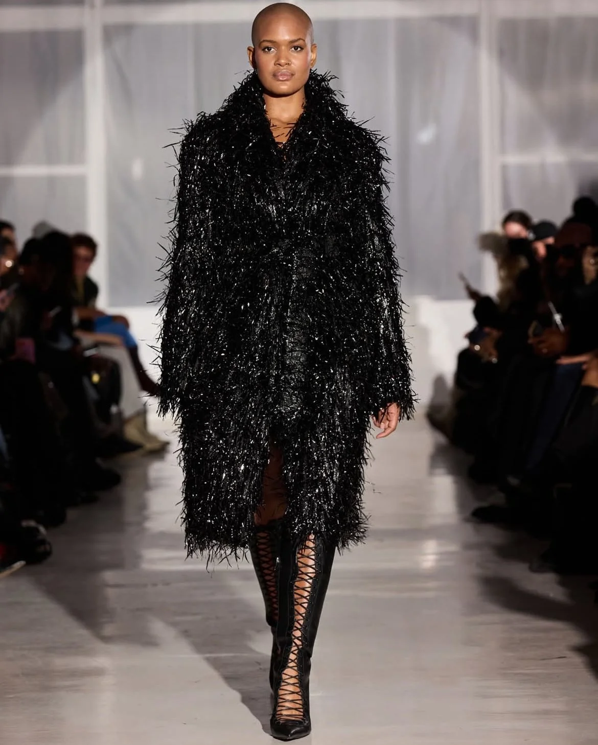 A model walks down a runway during a fashion show, wearing a long black textured coat and black lace-up high-heeled boots.
