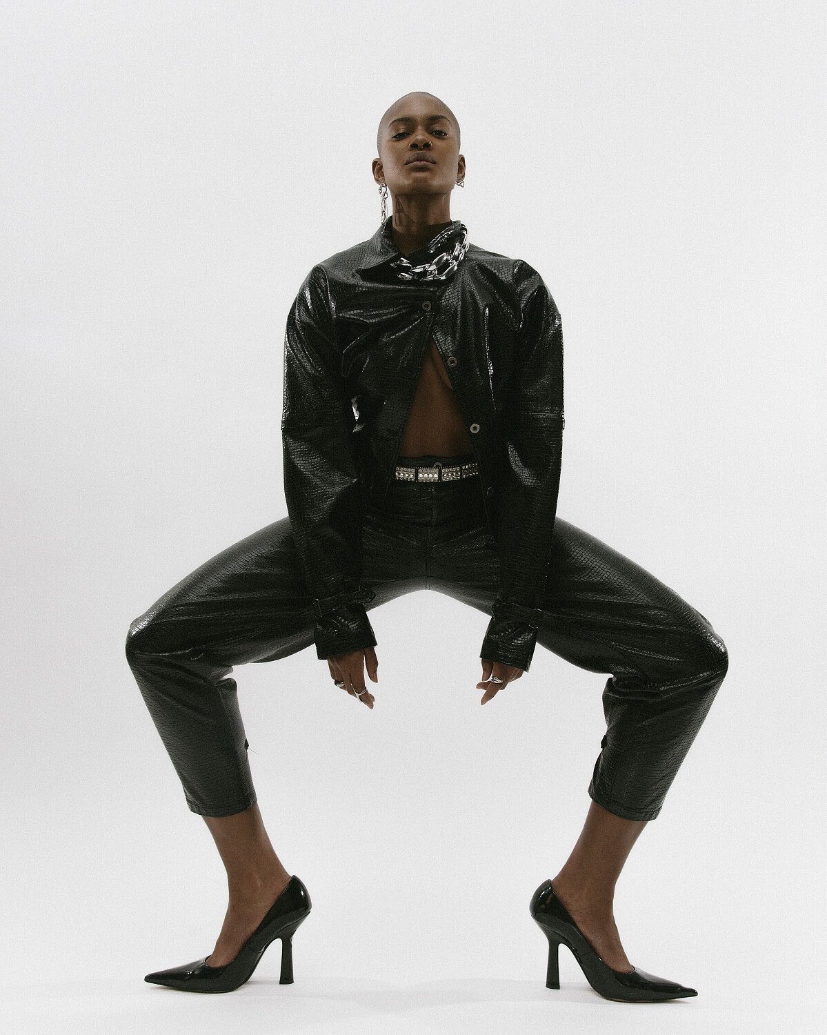 A person with a shaved head wearing a black shiny jacket, black shiny pants, black high heels, and jewelry, posing against a plain white background.