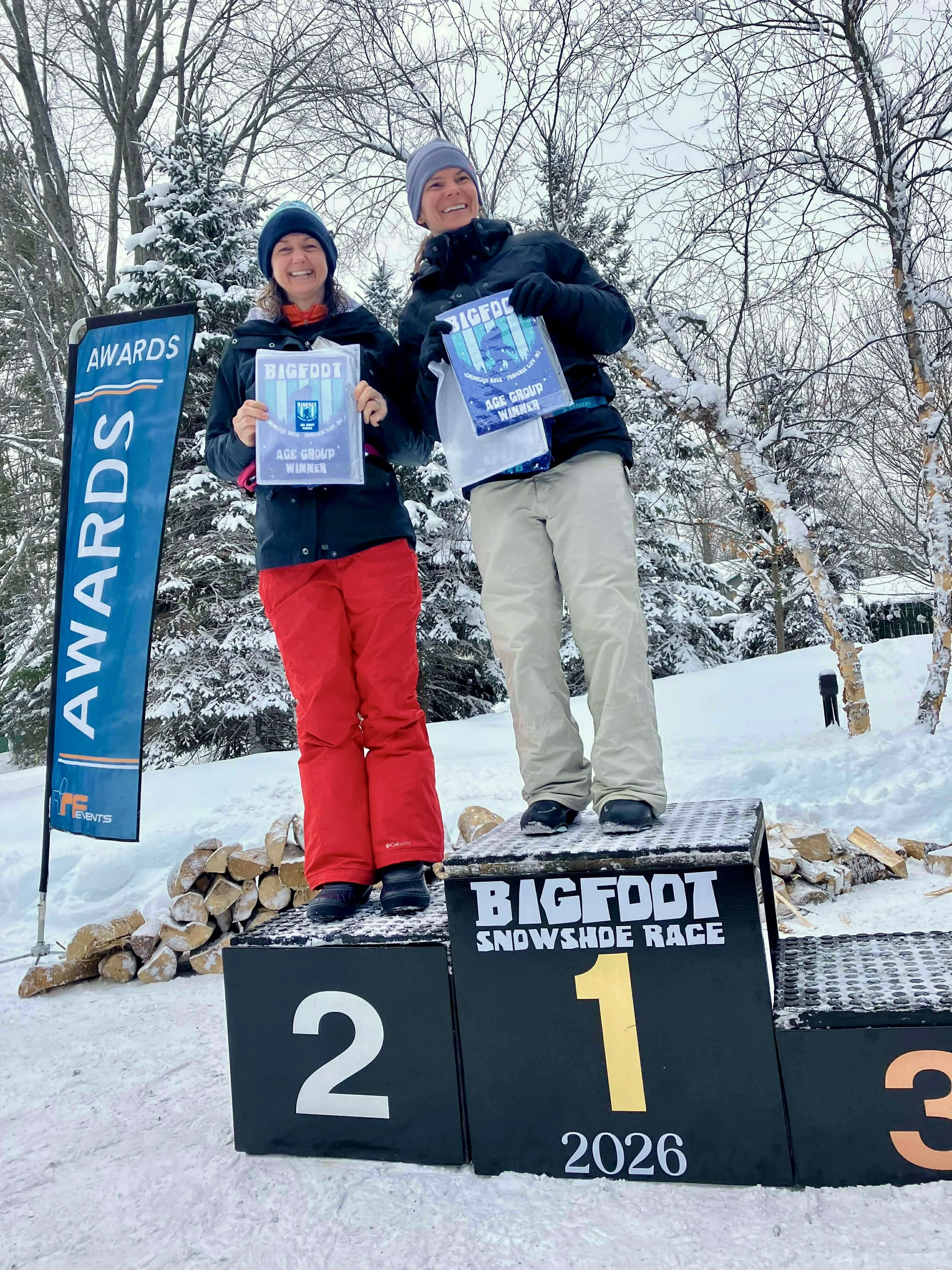 Two women standing on a winners' podium outdoors in a snow-covered landscape, holding awards at the Bigfoot Snowshoe Race. One woman is in first place, and the other is in second place. The background features snow-covered trees. A blue banner with the word 'AWARDS' is visible.