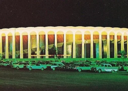 A line of vintage cars parked in front of a large, brightly lit building with colorful arches at night.