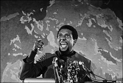 Man speaking at a podium, gesturing passionately, with a microphone setup in front of him and a large world map behind him.
