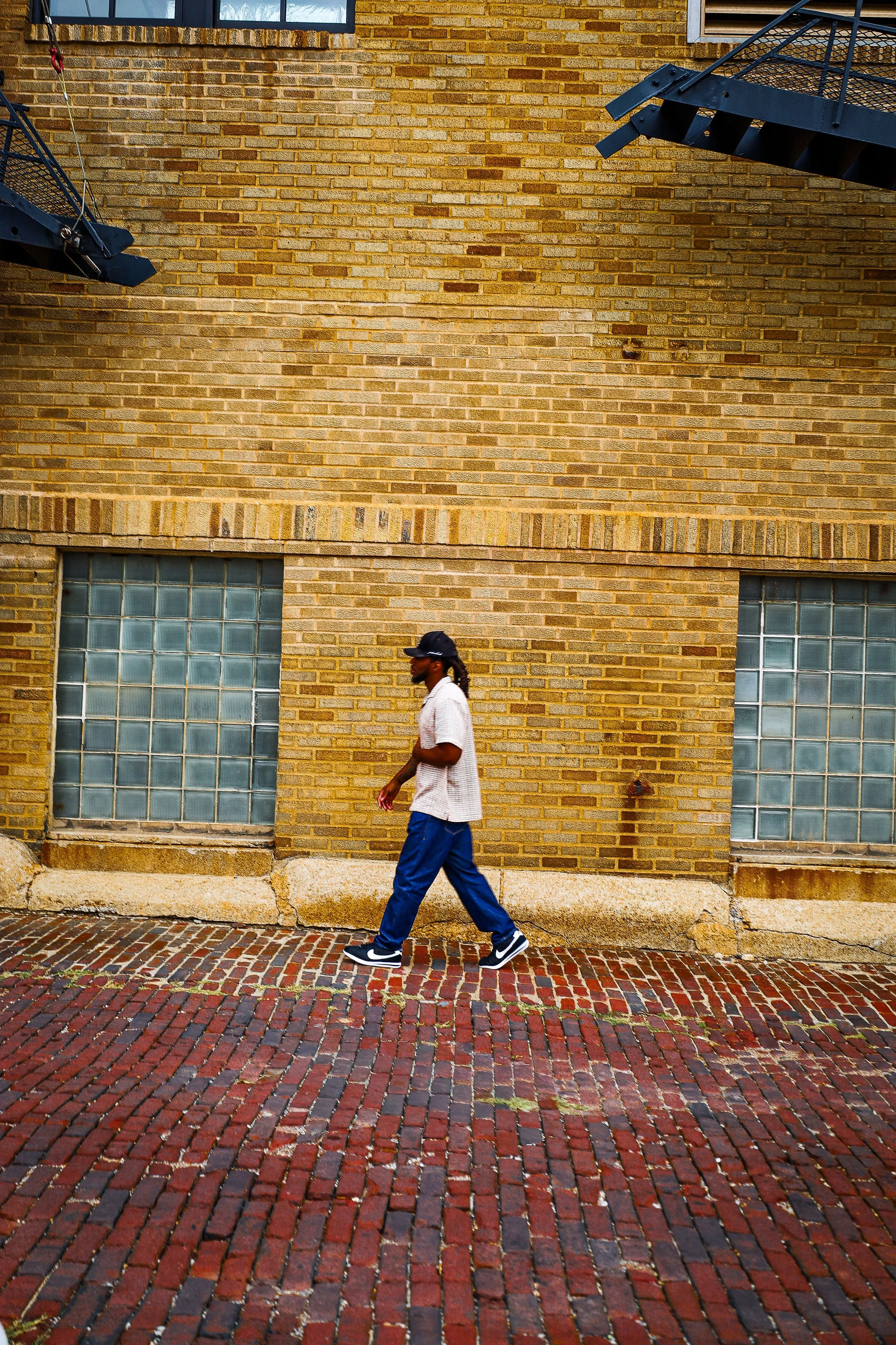 A man walking on a brick sidewalk in front of a yellow brick building with glass block windows. The man has long hair, wears a baseball cap, a white shirt, blue pants, and black sneakers.