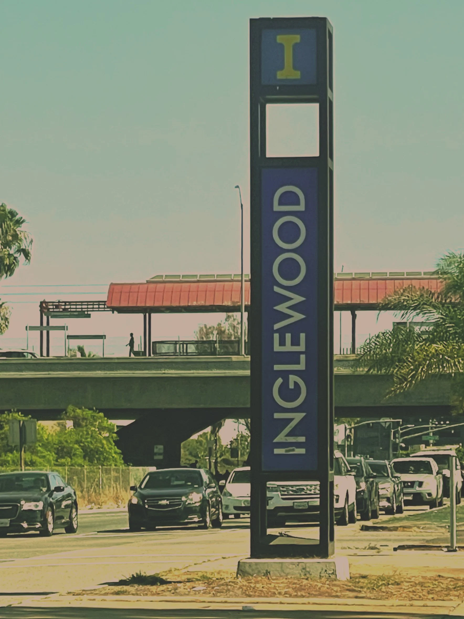 Parking lot with multiple cars parked and a vertical sign that reads "NO GAWED." In the background, an overpass with a red-tiled roof and some trees nearby.