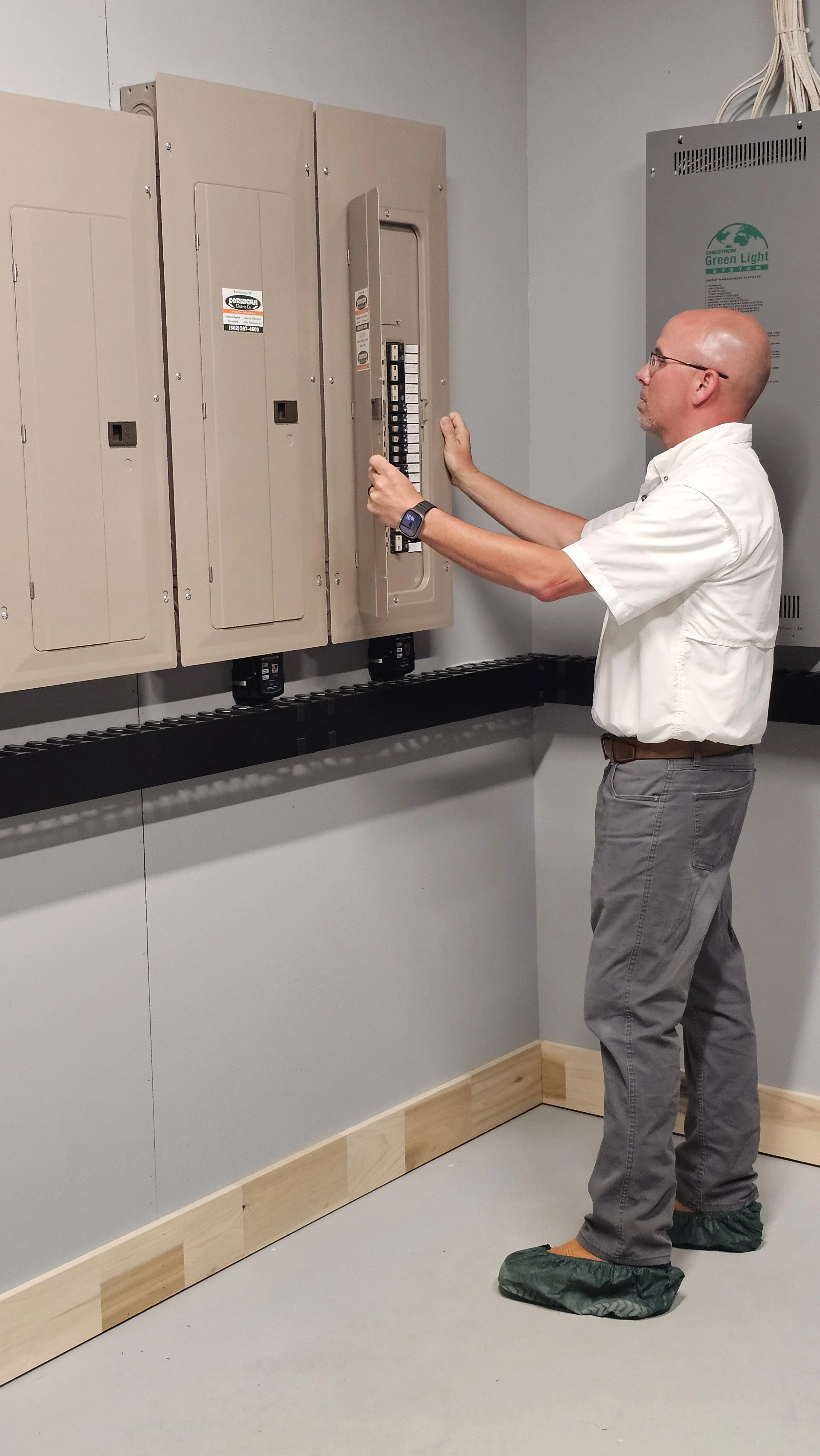 A man wearing glasses, a white shirt, gray pants, and shoe covers is working on electrical panels in a room with gray walls and a wood trim. He appears to be adjusting or inspecting the circuit breakers or switches within the panel.