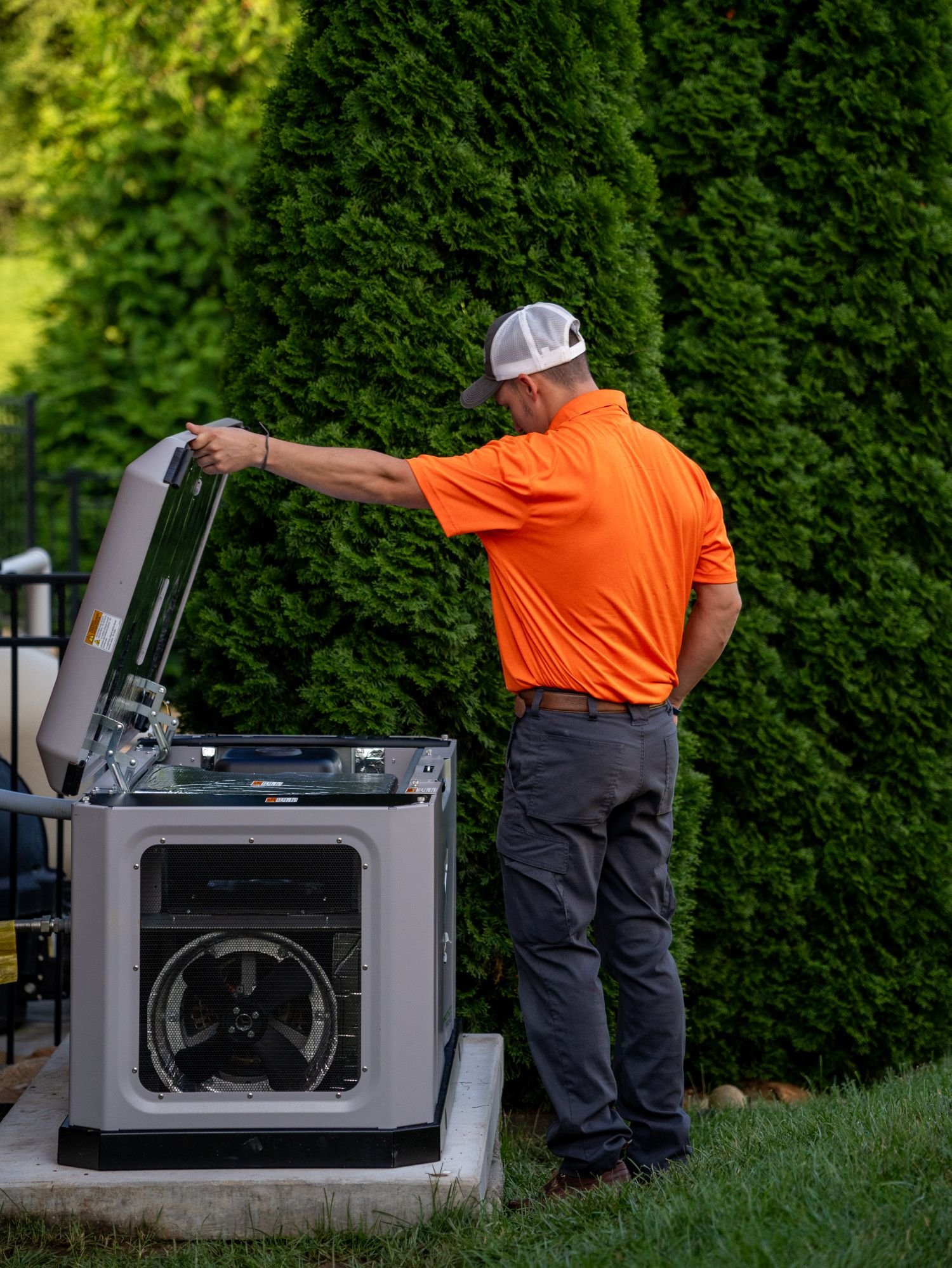 A man in an orange shirt and gray pants is standing outdoors near a HVAC unit, adjusting or inspecting it with greenery in the background.