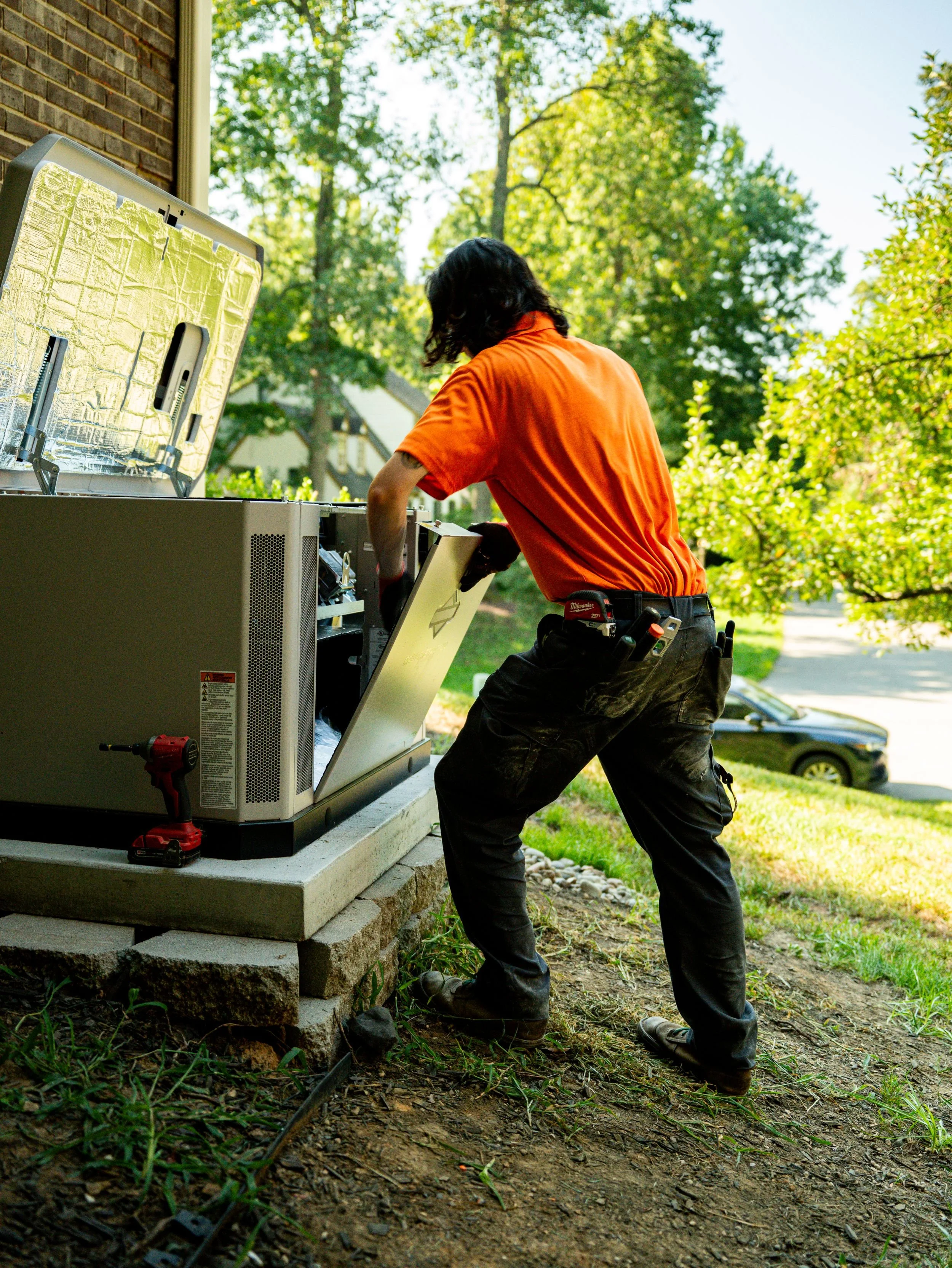 A technician installing or repairing an electrical or HVAC unit outside a house on a concrete platform surrounded by a grassy yard, with trees and cars in the background.