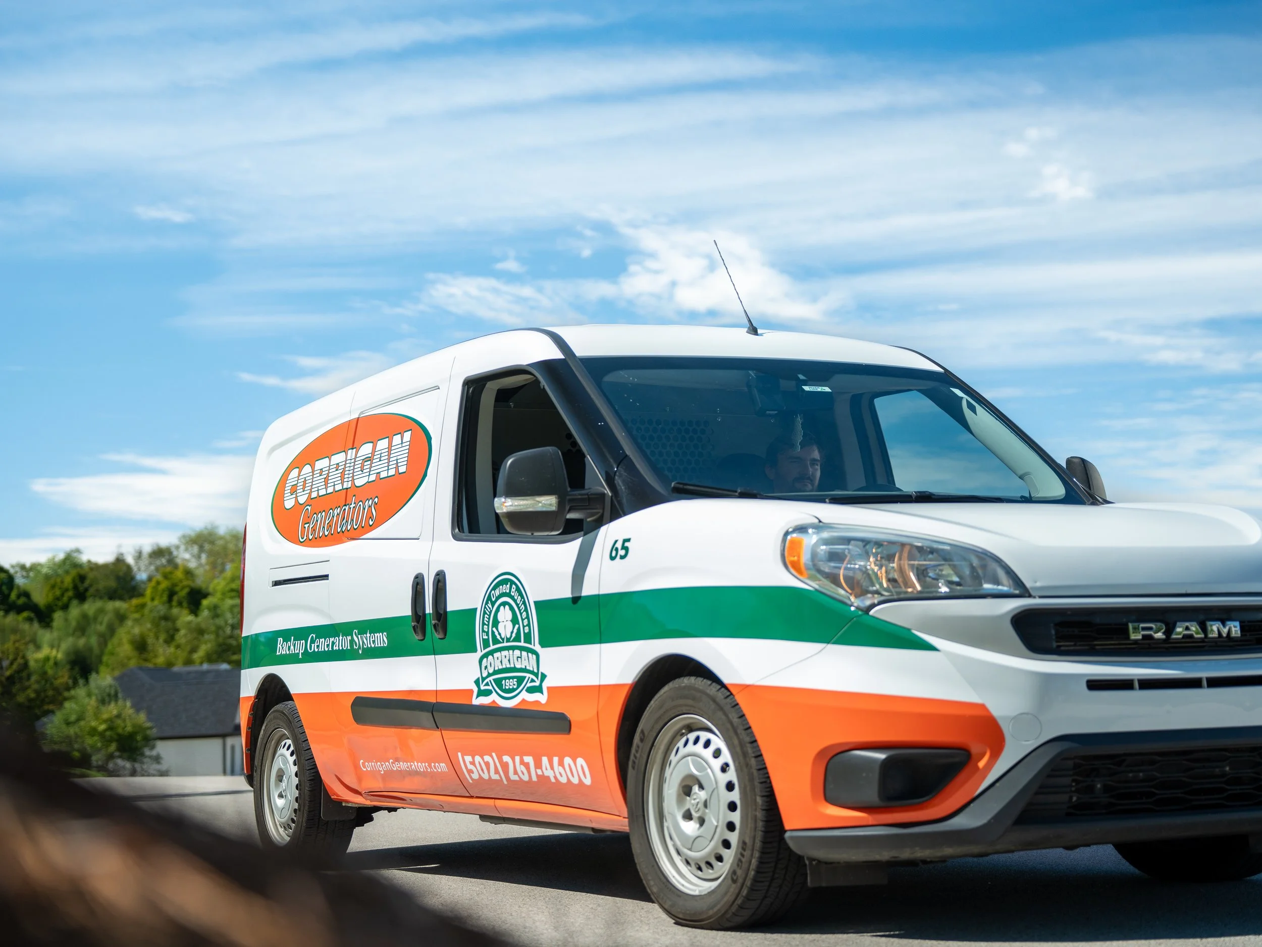 A white company van with orange and green branding for Corrigan Generators is driving on a road under blue skies with some clouds. The van has contact information and a logo on its side.