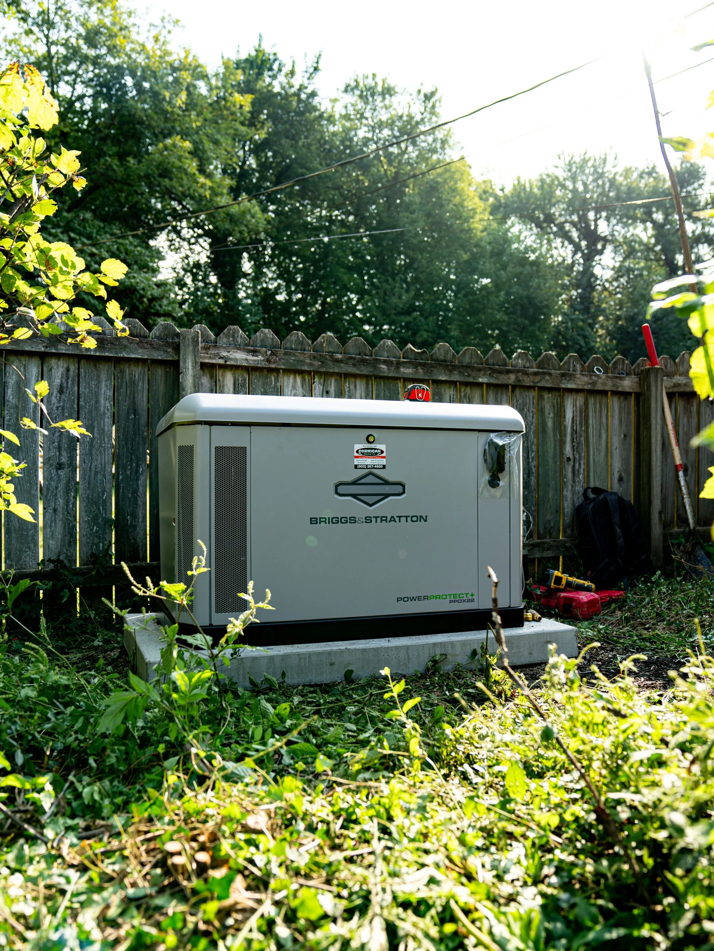 An outdoor scene with a fenced backyard, a gray electrical generator on a concrete pad, and tools and a backpack nearby, surrounded by green plants and trees, with sunlight shining through.