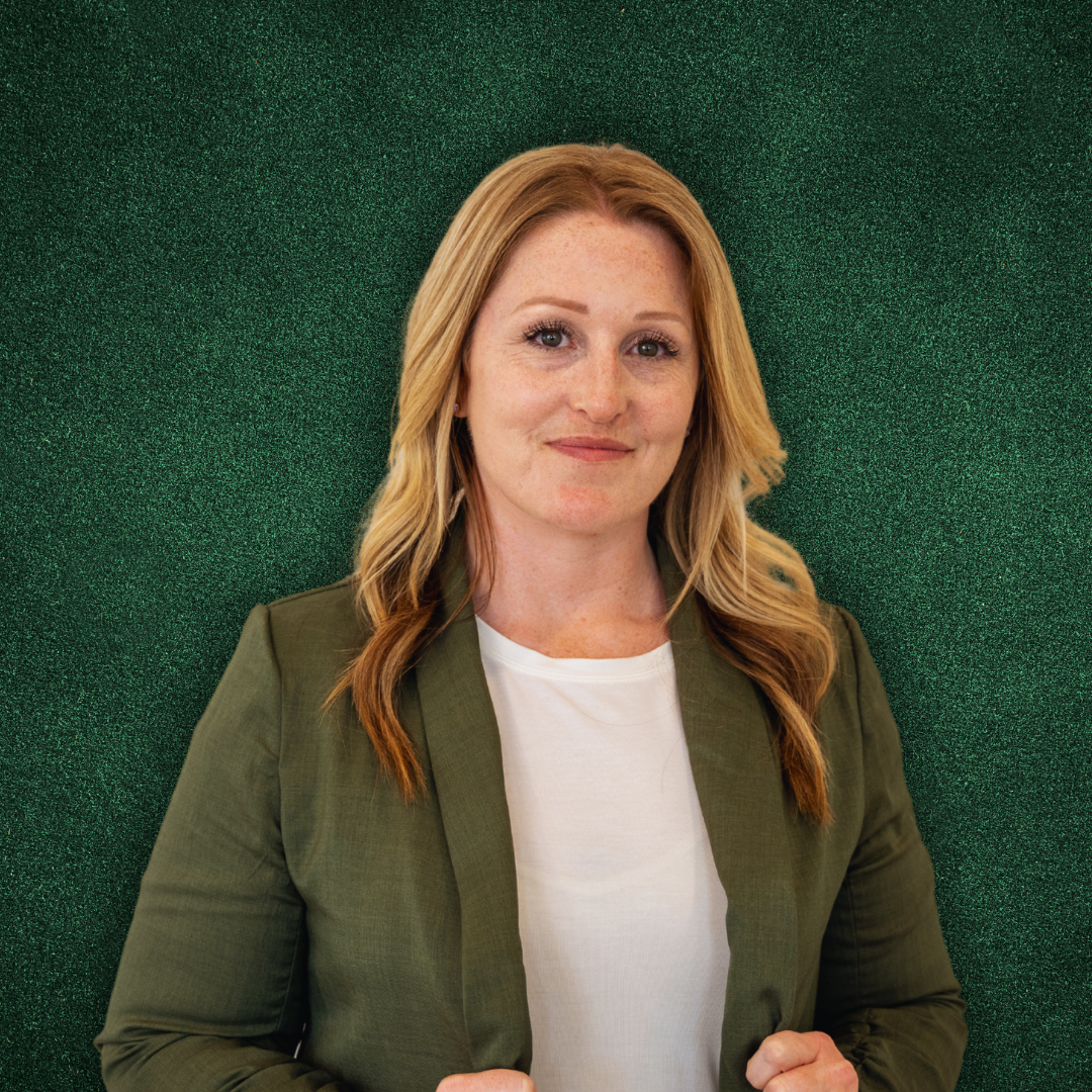 A woman with long wavy red hair, wearing a white t-shirt and an olive green blazer, standing in front of a textured green background.