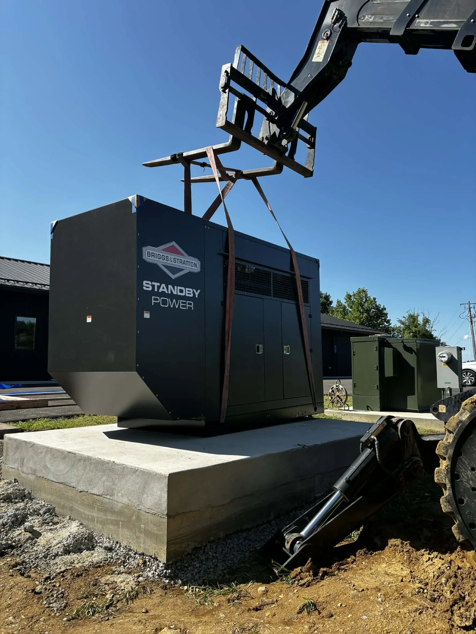 A large portable power generator with the label 'Briggs & Stratton Standby Power' sits on a concrete pad outside a building. A construction lift with a platform is extended upward, likely being used to install or maintain the generator. The sky is clear and blue, and there are trees and other structures in the background.