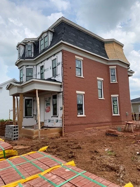 A three-story brick building under construction with visible framing and roofing work, surrounded by construction materials and dirt ground.