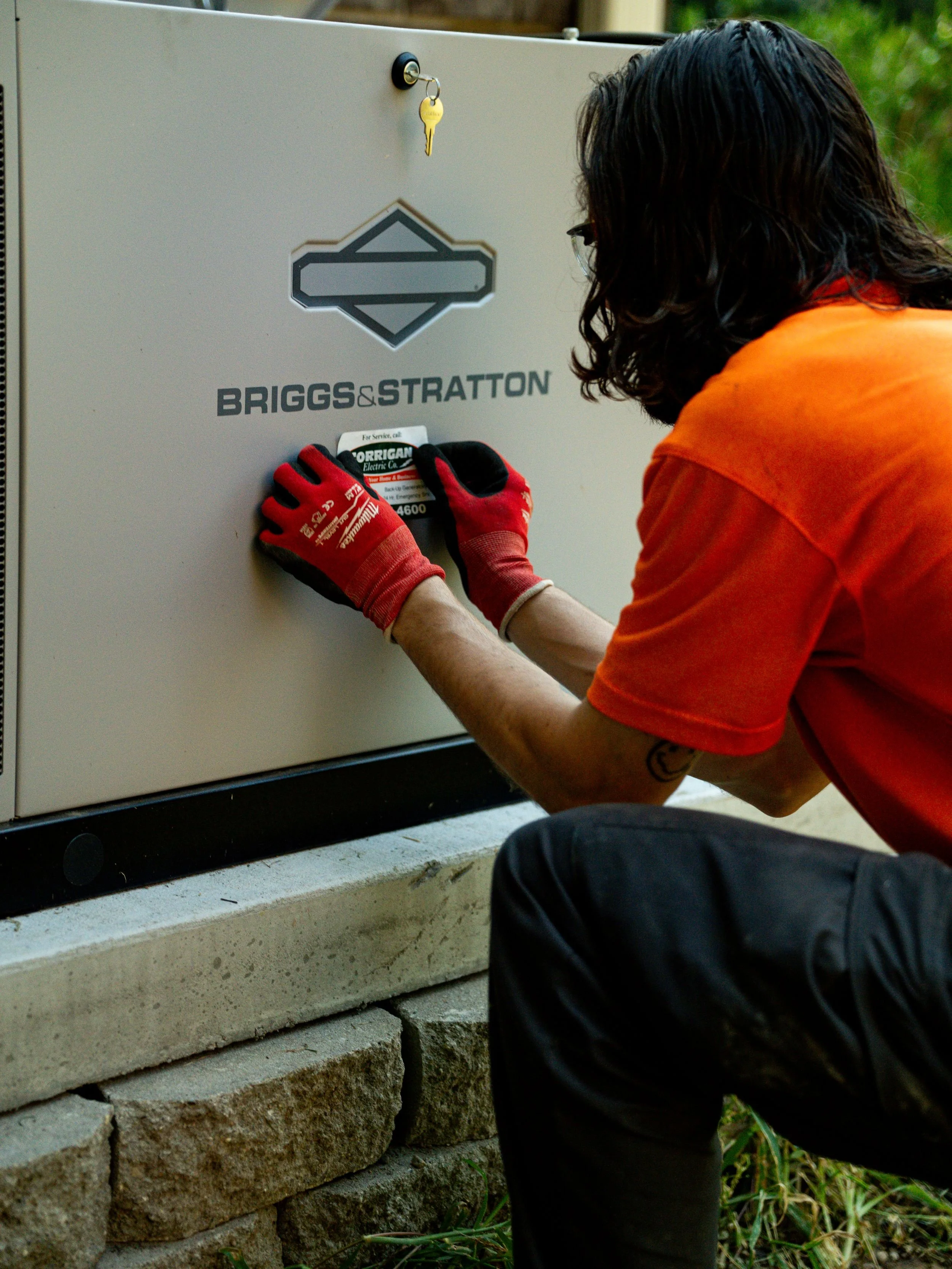 A person wearing red gloves and glasses is installing a sticker with a black and white logo and text on a gray electrical box, with stacked concrete blocks underneath and greenery in the background.