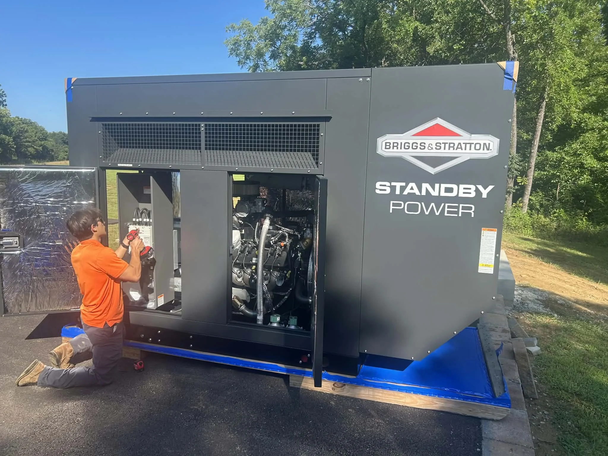 A child kneeling and working on a large industrial generator with the label "Briggs & Stratton Standby Power." The generator is outdoors, surrounded by trees and a clear blue sky.