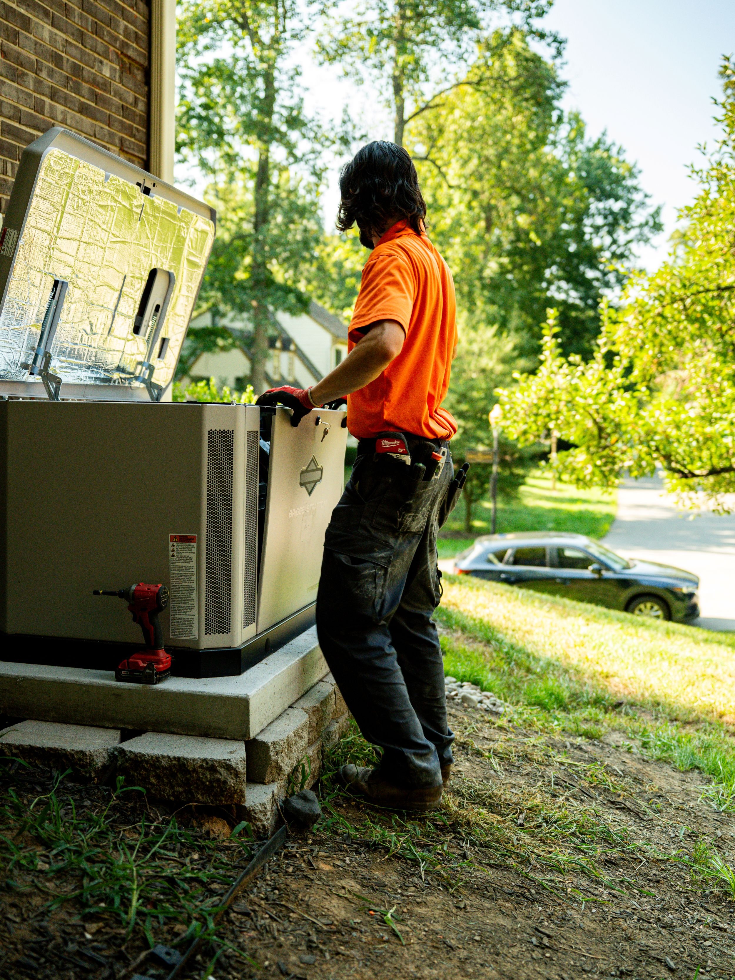 A technician with long dark hair, wearing an orange shirt, black work pants, and gloves, working on an outdoor HVAC unit on a concrete slab, with tools attached to their belt, in a residential area with trees and a car in the background.