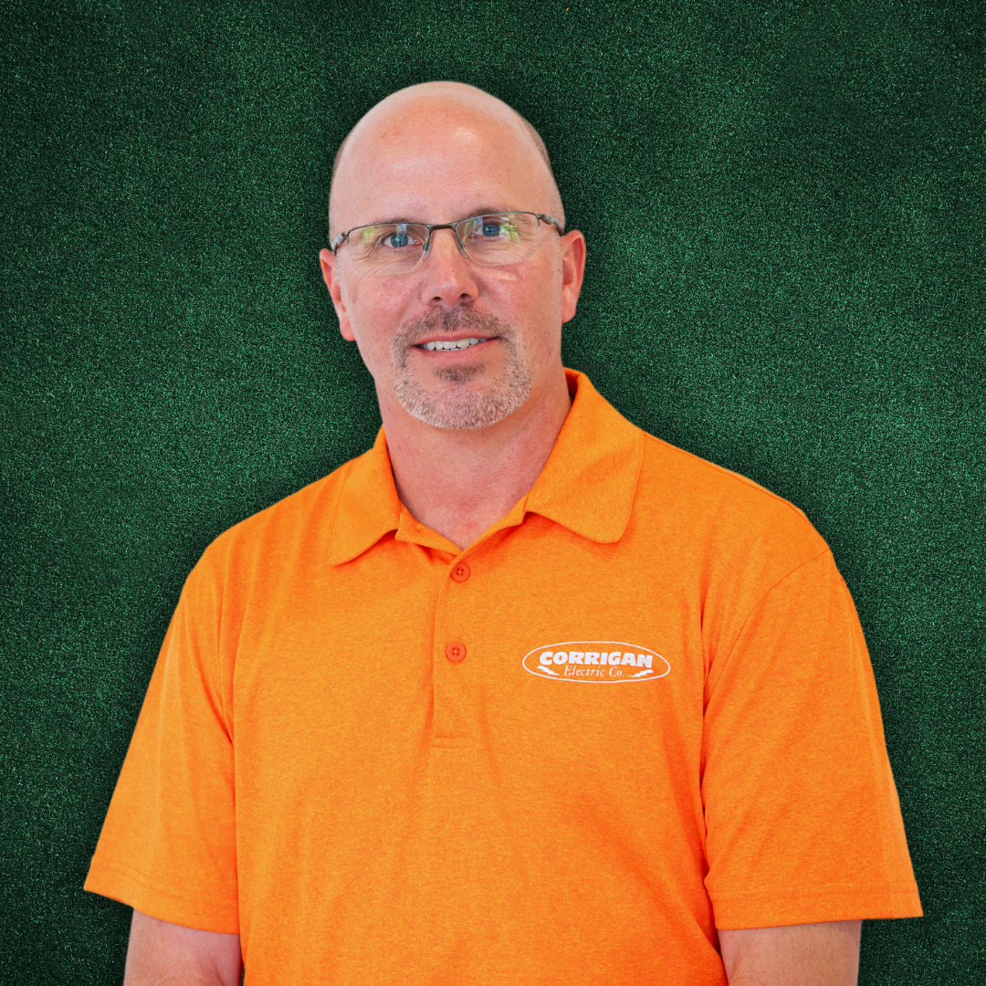 A man with glasses and a goatee wearing an orange polo shirt with a logo that says 'Corrigan Electric Co.' stands in front of a green background.
