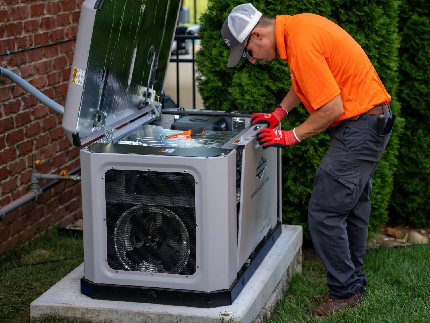 A technician wearing an orange shirt, gray pants, red gloves, and a baseball cap repairing or maintaining a solar-powered generator outside near a brick wall and green bush.