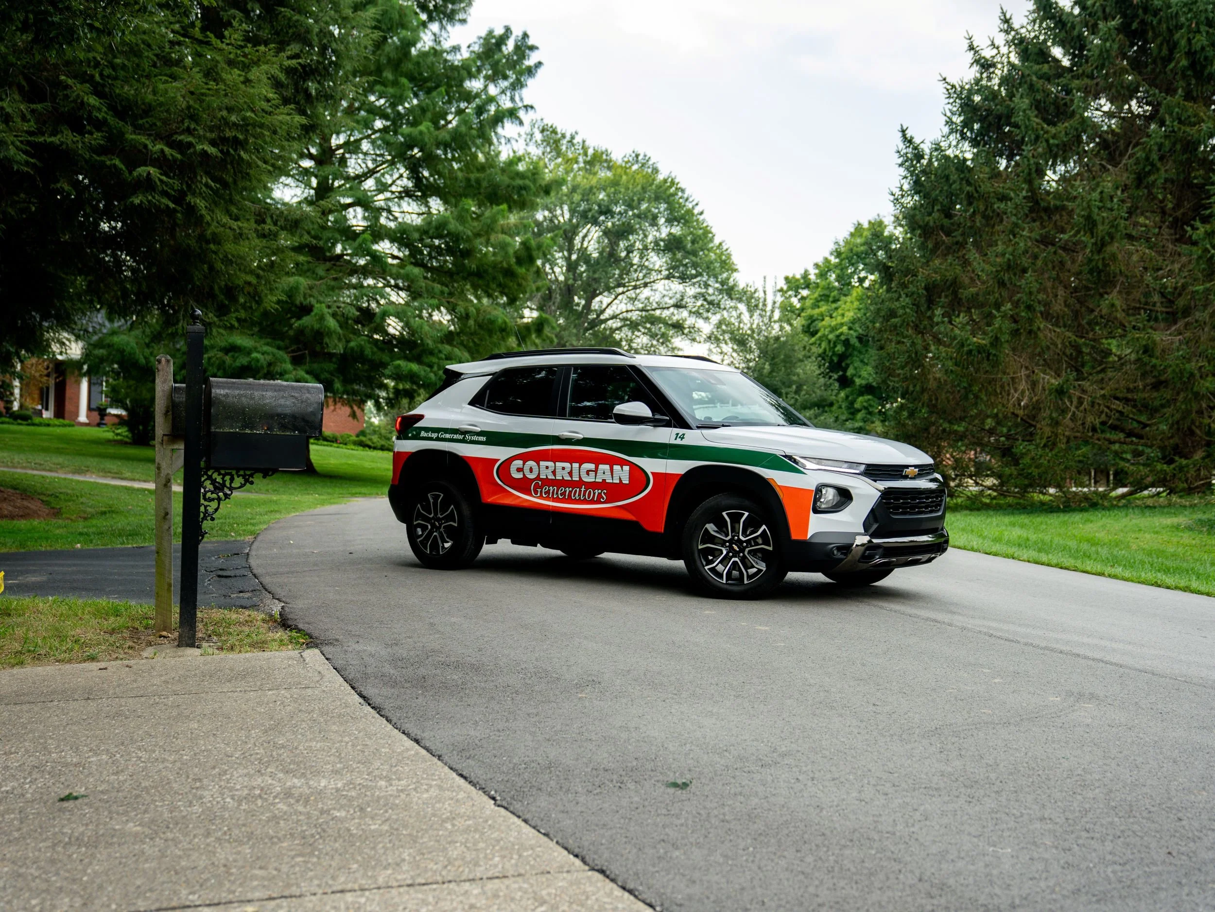 A white Chevrolet SUV with red, green, and black markings for Corrigan Generators parked on a curved driveway surrounded by green trees and a grass lawn.