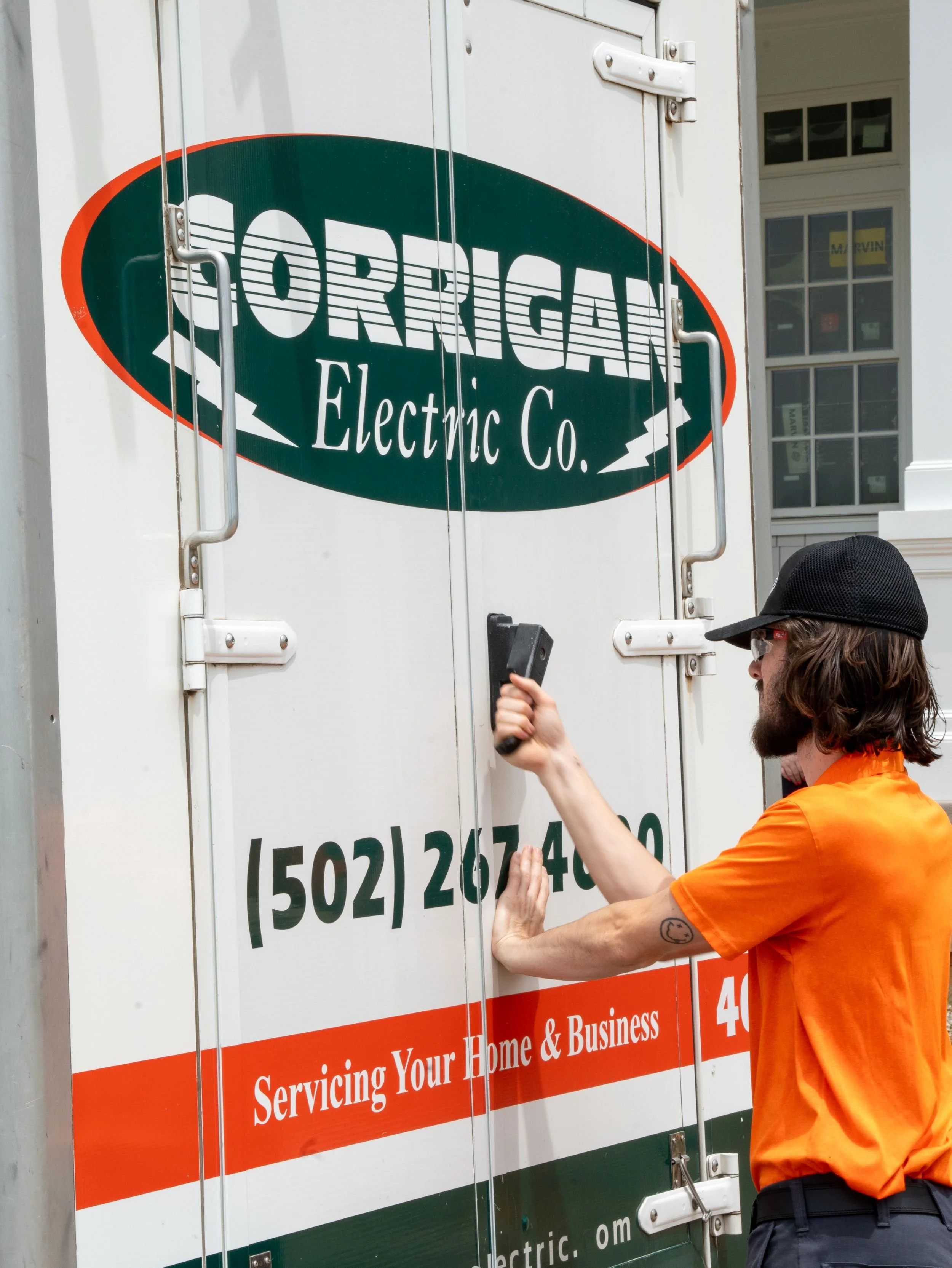 A person with long hair, glasses, and a beard, dressed in an orange shirt and a black cap, is opening a white trailer with a logo for 'Orrigan Electric Co.' on it. The trailer has red and green signage.
